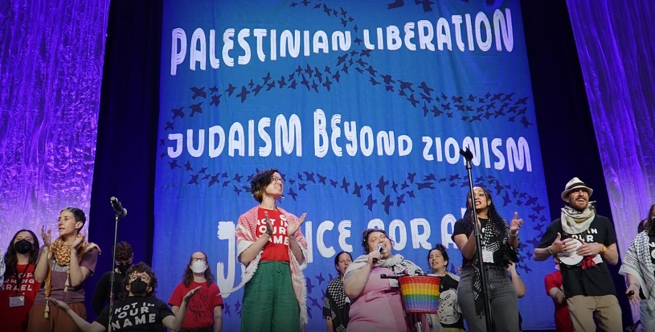 Diverse crowd of people clapping on stage. Giant banner behind them reading " PALESTINIAN LIBERATION - JUDAISM BEYOND ZIONISM -- JUSTICE FOR ALL"