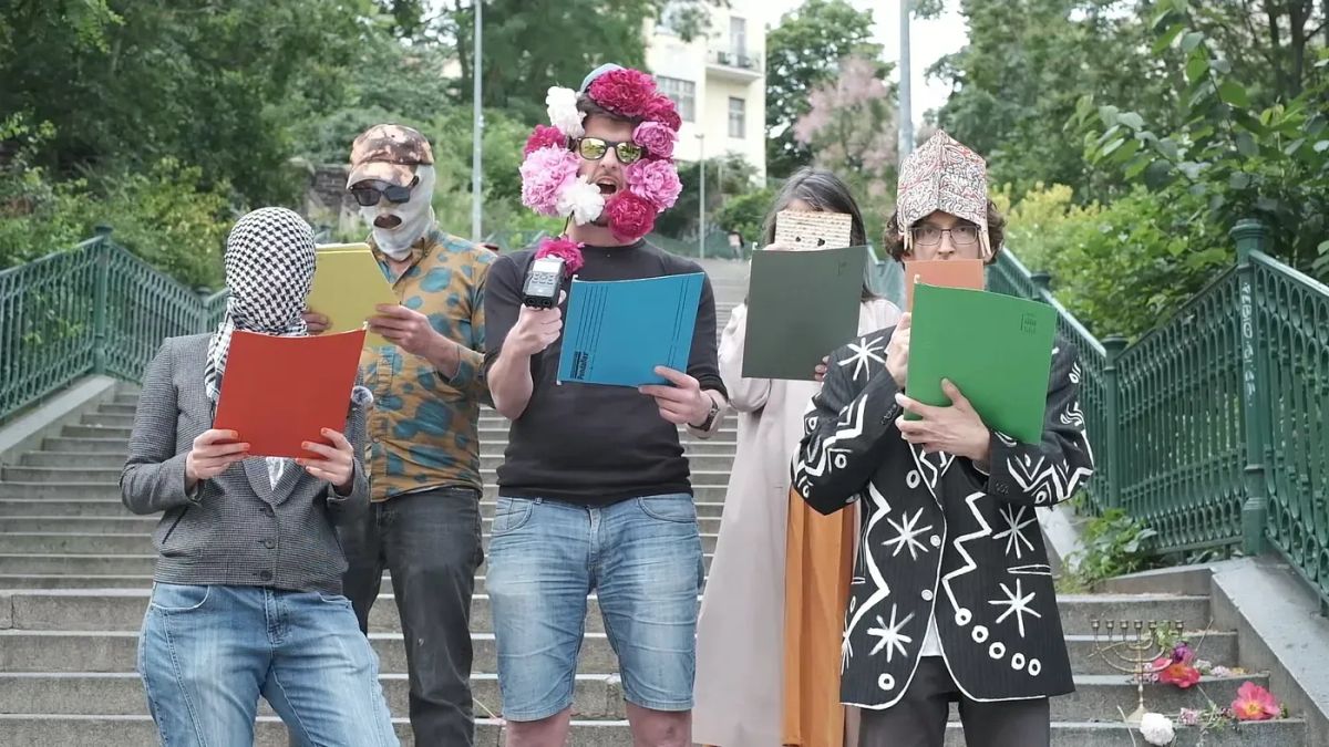 Several people masked in flowers a keffiyeh, and matzoh read from the declaration of independence of diaspora in a park.