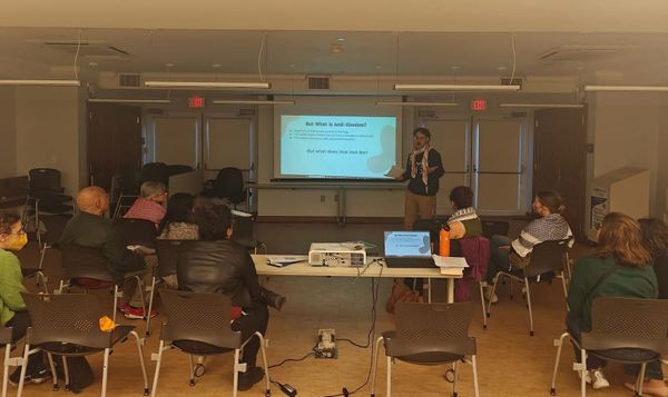 A person in a keffiyeh speaks to a group of 9 people while standing in front of a slideshow on a projected screen.