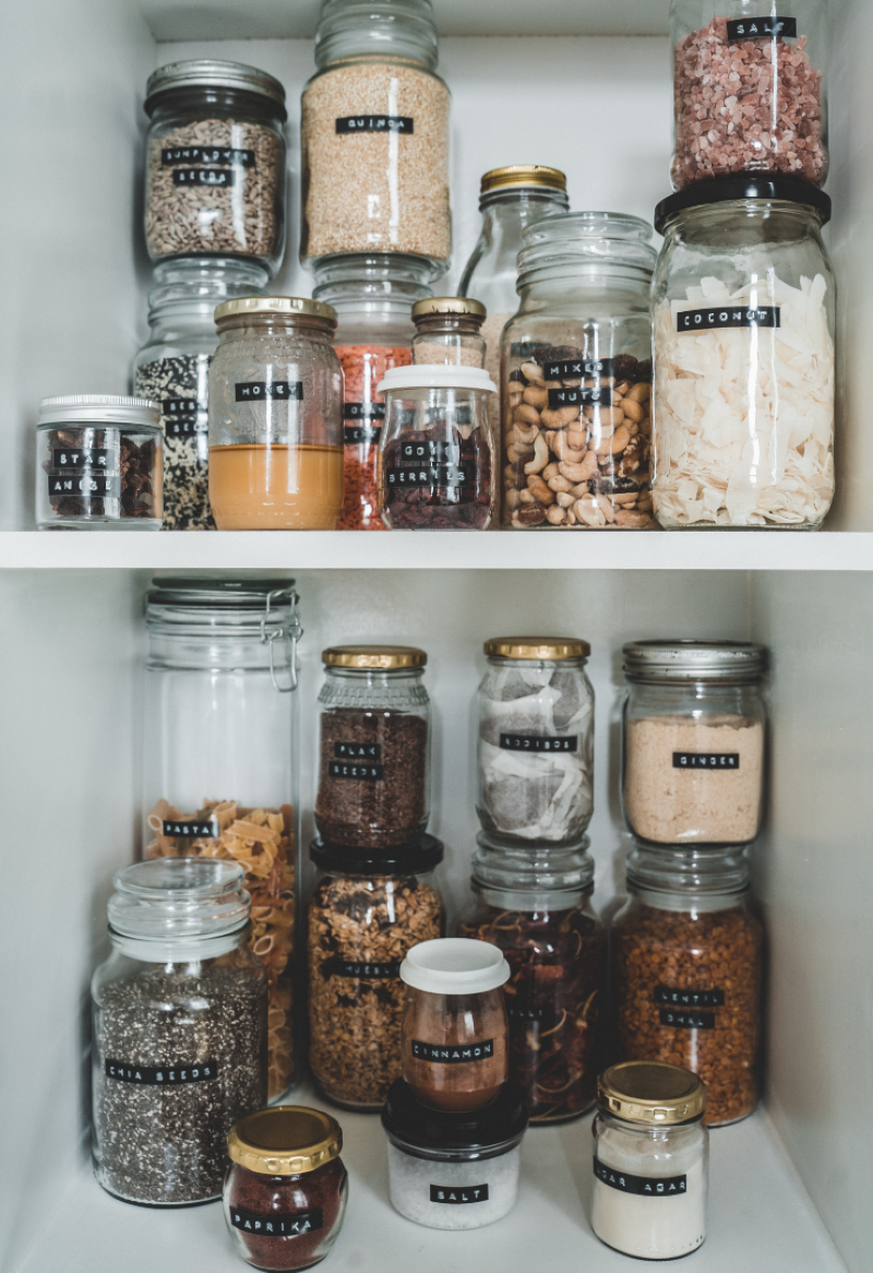 Barattoli di vetro etichettati organizzano una dispensa piena di ingredienti secchi, tra cereali, semi, spezie e frutta secca.