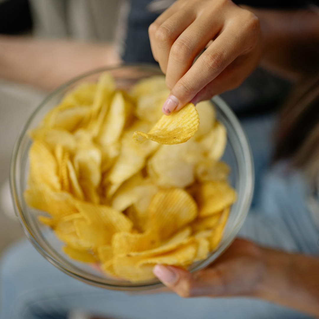 Mano che prende una patatina da una ciotola trasparente piena di snack, immagine simbolica per una riflessione sulla comunicazione alimentare, sui gesti di consumo e sulla responsabilità dell