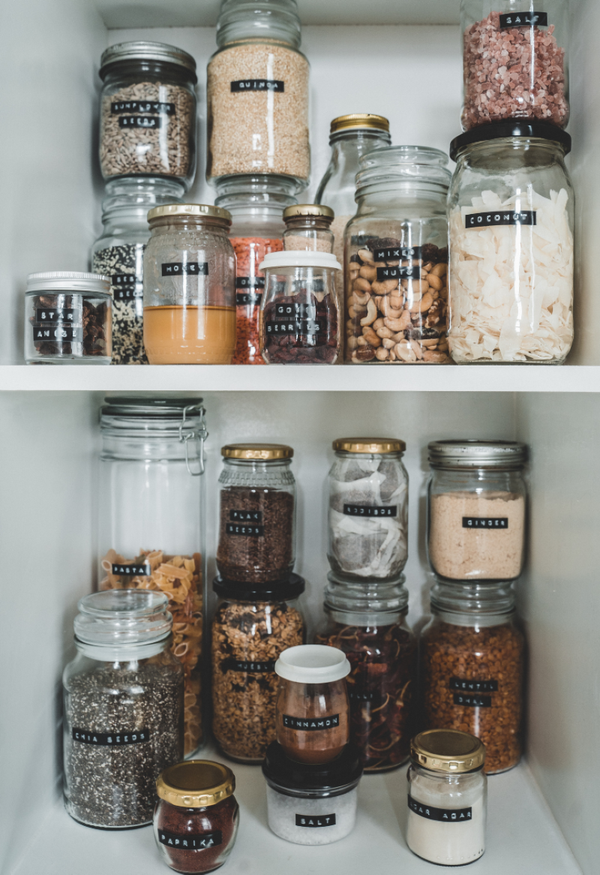 Barattoli di vetro etichettati organizzano una dispensa piena di ingredienti secchi, tra cereali, semi, spezie e frutta secca.