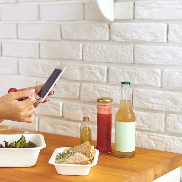 Mani che usano uno smartphone accanto a un pasto confezionato con panini, insalata e bevande in bottiglia, disposto su un tavolo di legno.