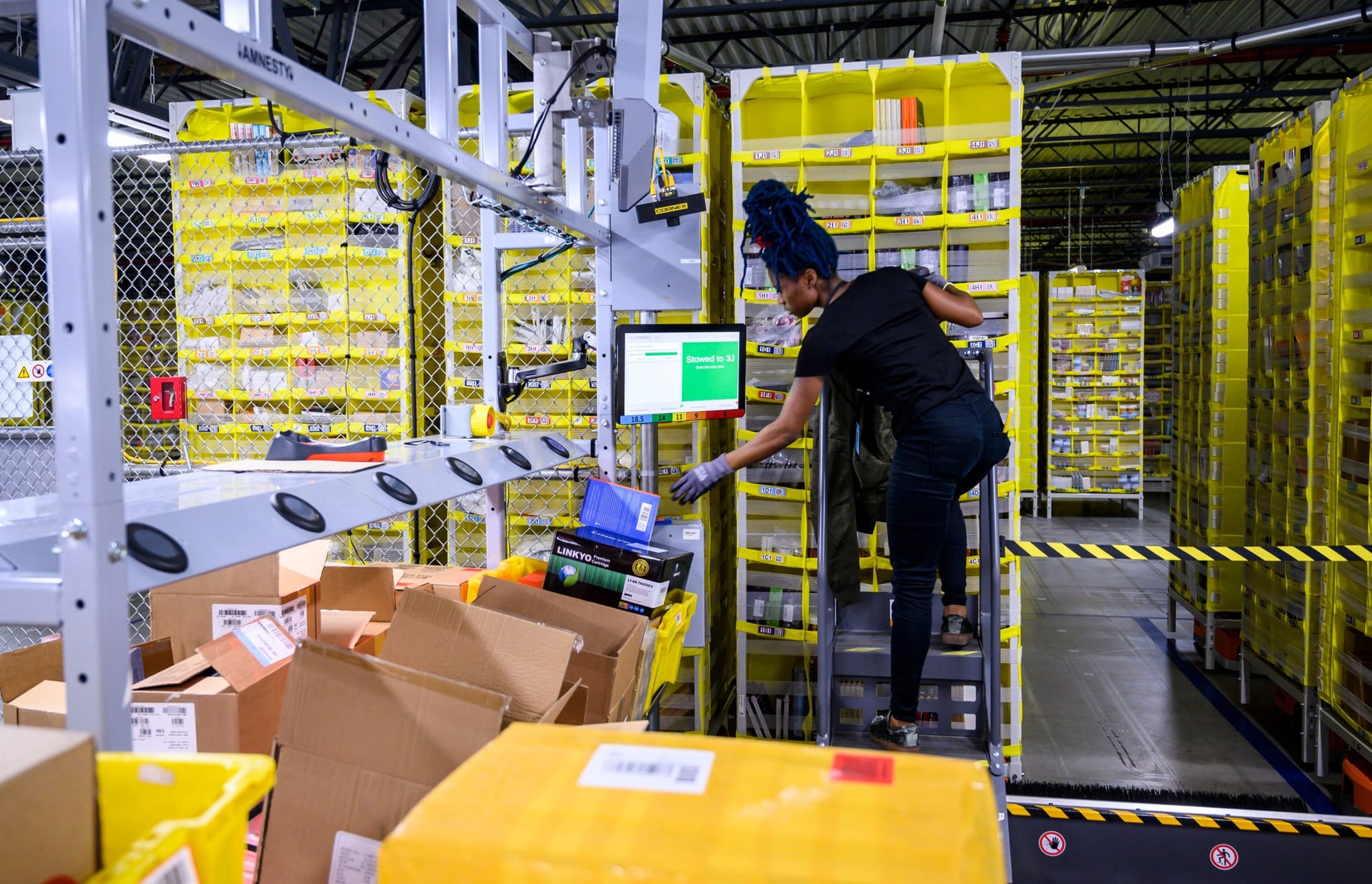 color photograph of a black woman standing on a metal step ladder surrounded by yellow shelves of items in an Amazon warehous