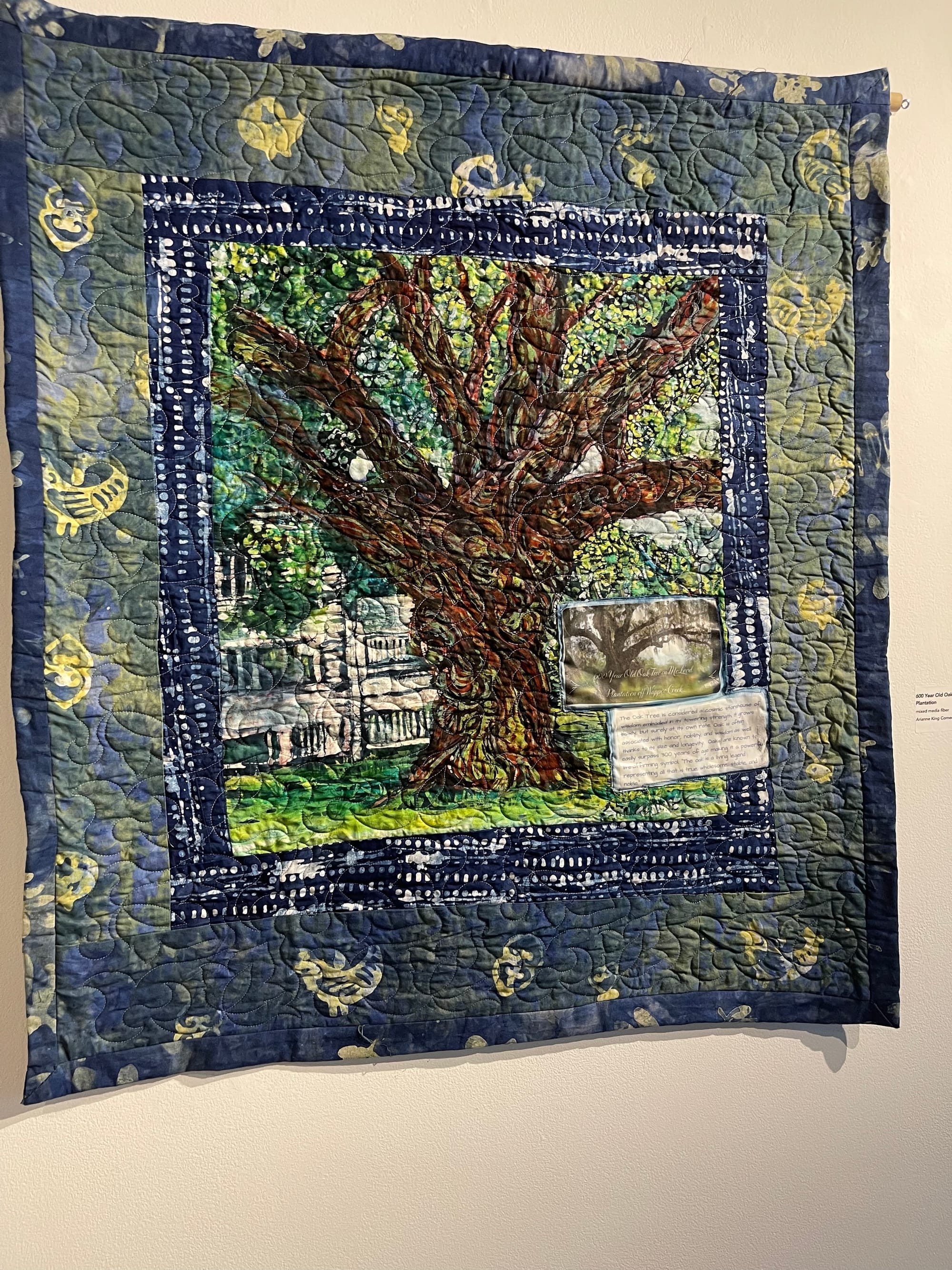 quilt hanging on an off-white wall. the quilt has a ring of blue with yellow crescents like stars, and in the center is a tree in front of a large white plantation house