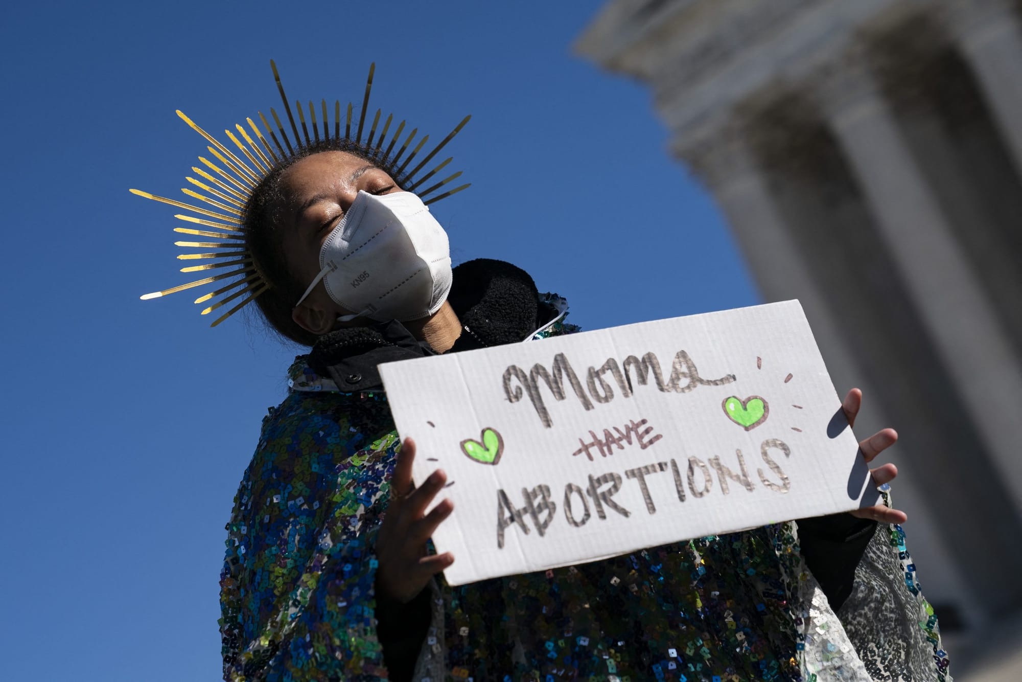 person wearing a sunlight tiara stands with their eyes closed hodling a sign reading "moms have abortions"