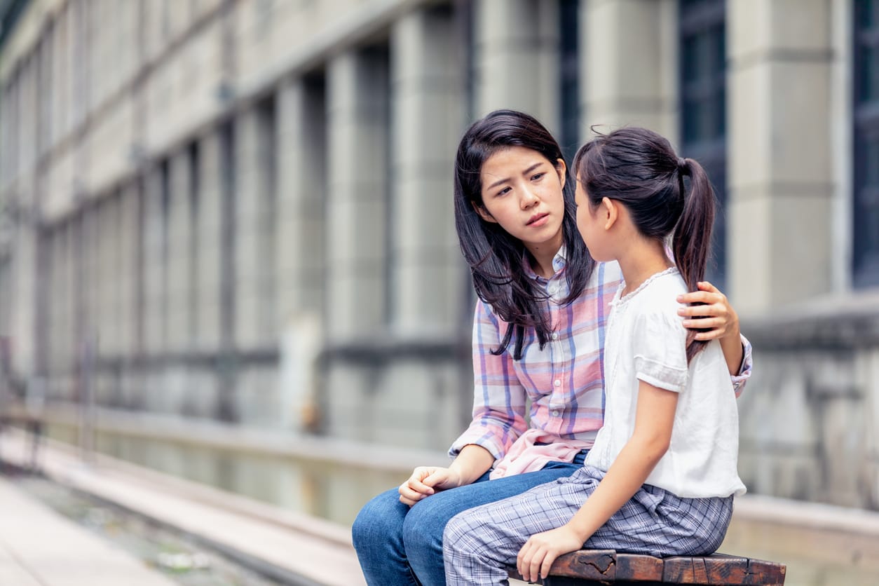 A young asian woman with dark hair sits with her arm around her child, looking serious. The child faces away from the camera.