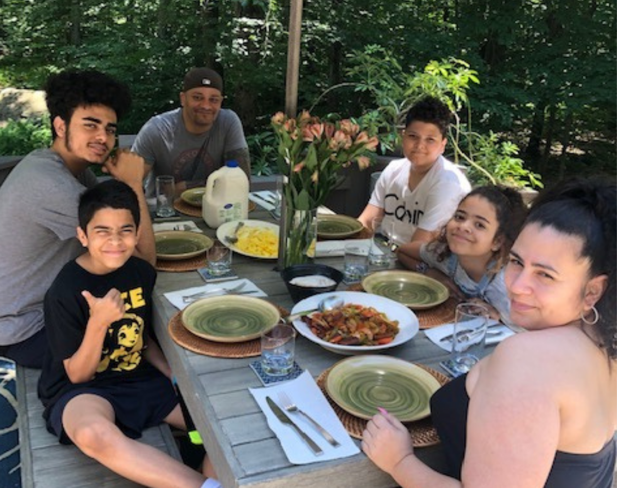 a latina mother sits closest to the camera with her family in the background. all of them are sitting at an outdoor table with food
