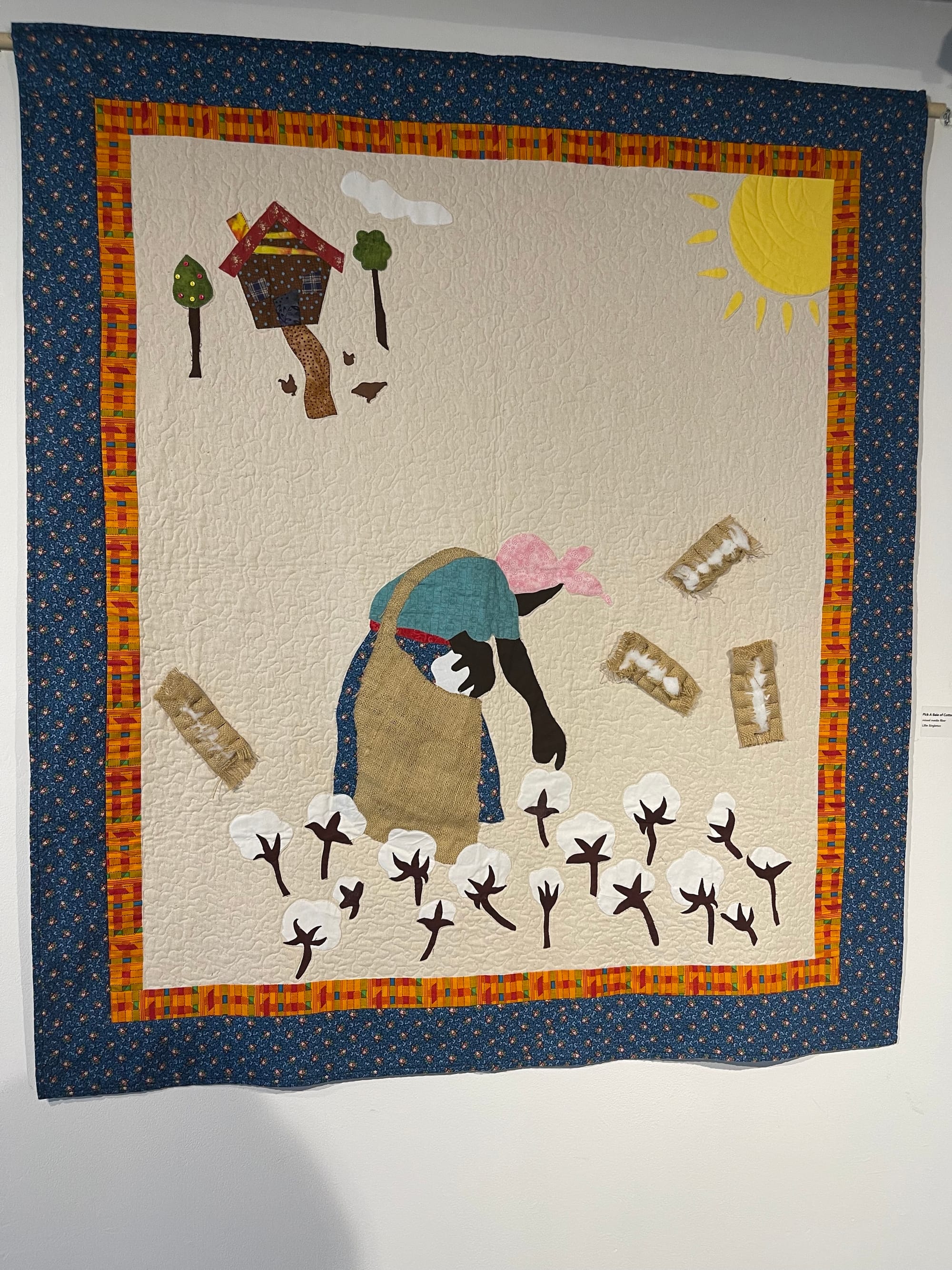 quilt hanging on an off-white wall. the quilt depicts a black woman wearing a pink head scarf and blue dresspicking cotton in front of a house in the background