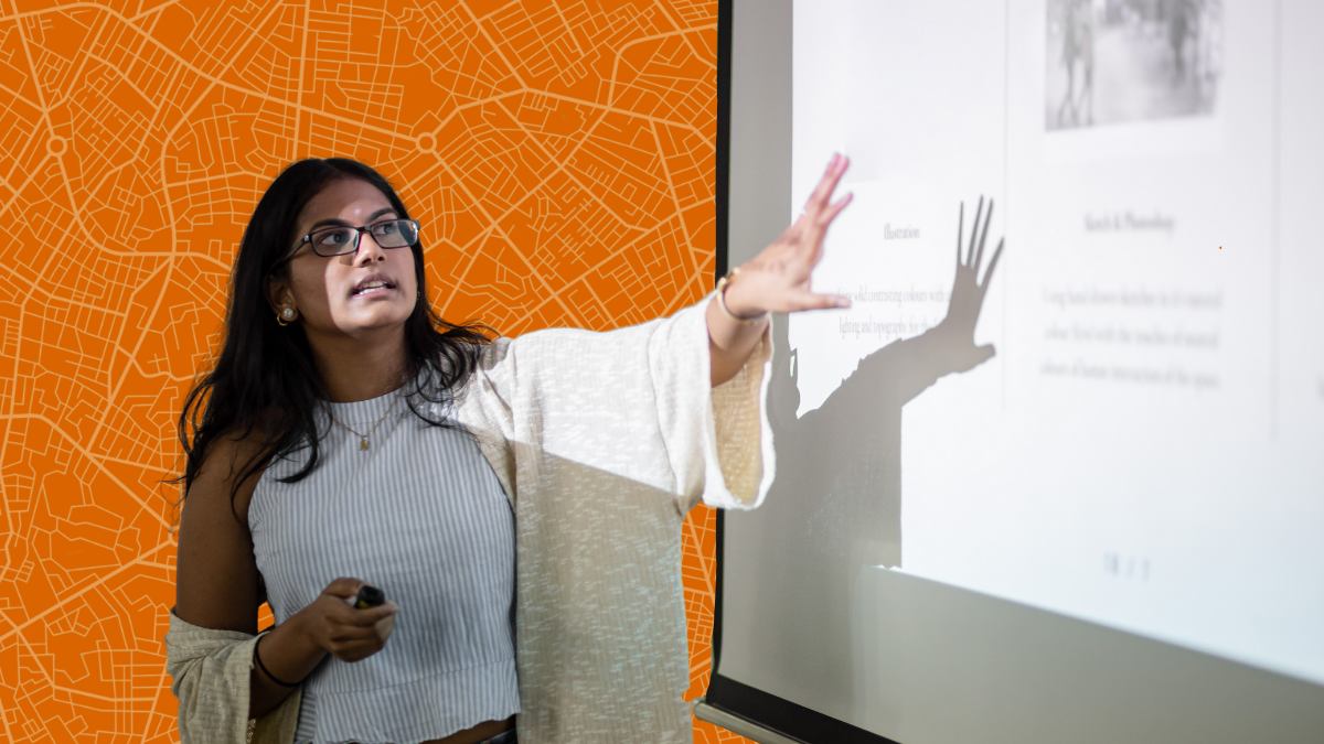 a woman with dark hair past her shoulders stands in front of a projecter screen and points towards it