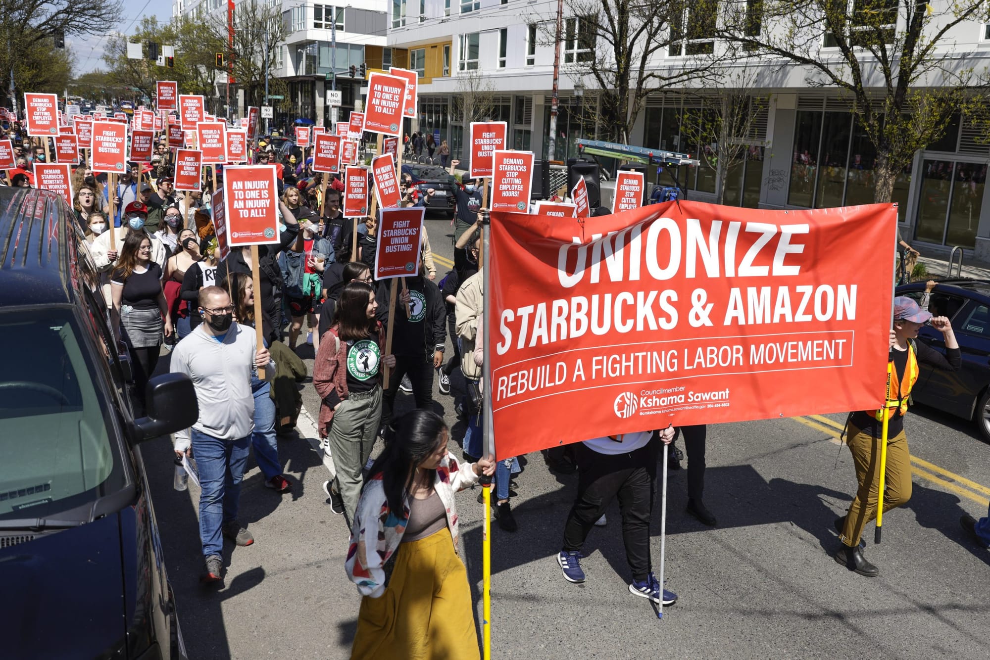 protesters hold a red banner reading "Unionize Starbucks and Amazon" as they march down a street lined with businesses and ap