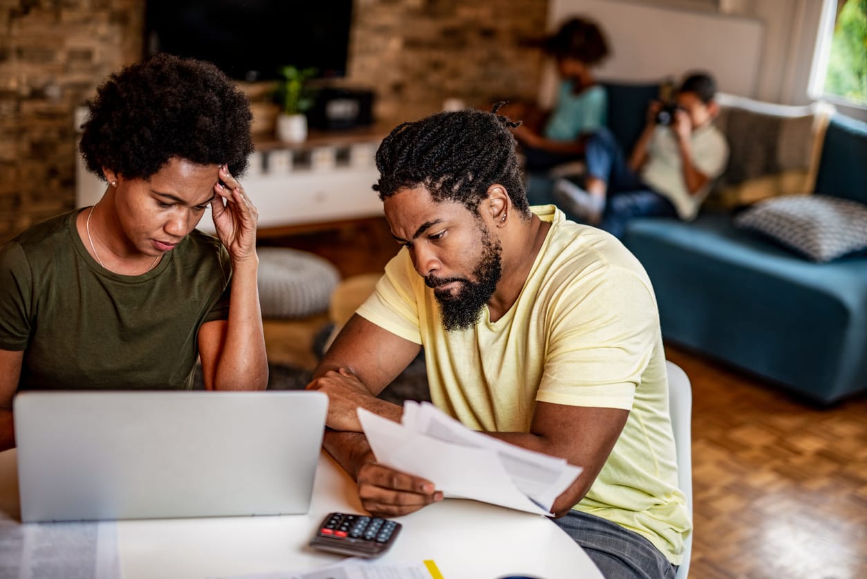 a Black couple sits at a table with a laptop, calculator, and papers spread out. they look stressed while their kids sit on t