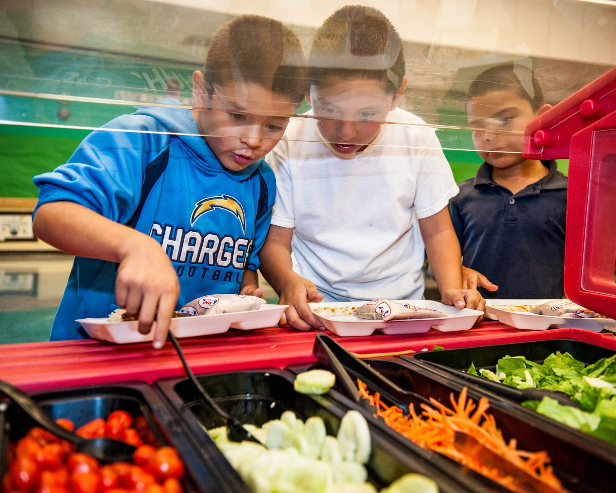 three elementary school-age boys line up in front of a salad bar holding black plastic spoons to pick up lettuce, carrots, an