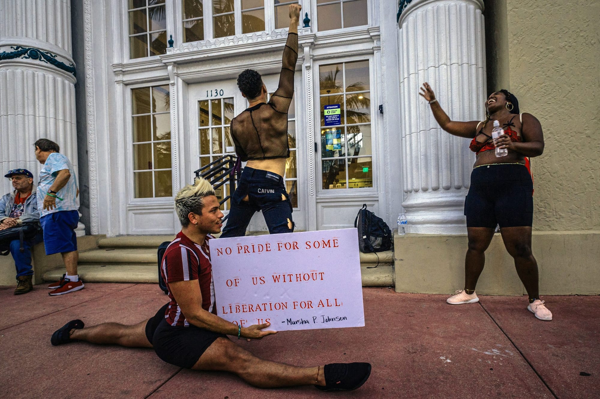 a person with bleached hair does the splits parallel to the camera while holding a white sign with red text reading "No pride
