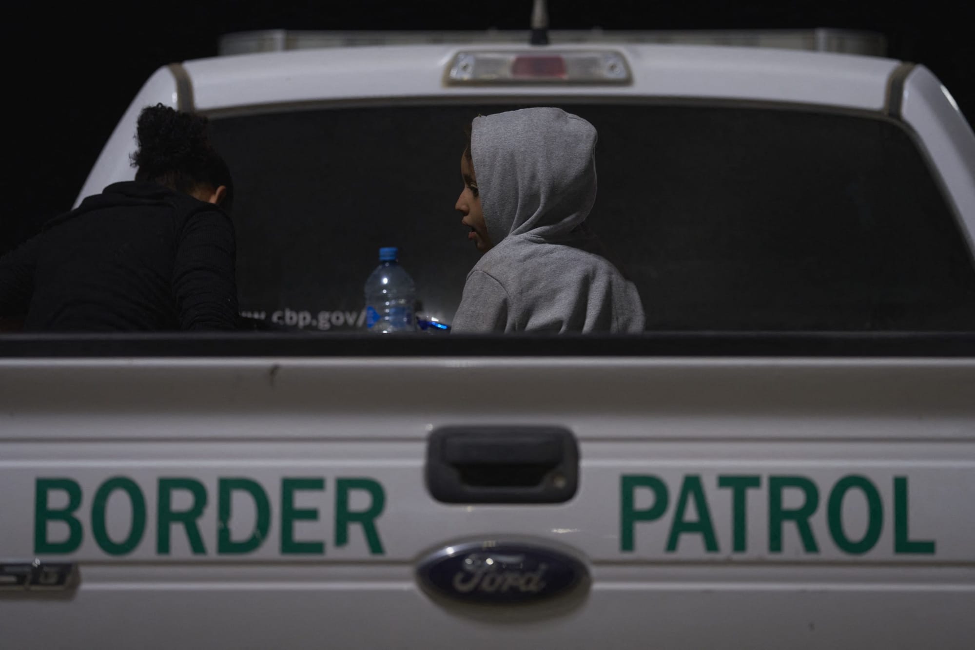 a child wearing a grey hoodie with the hood up sits in the bed of a white pick-up truck with the words "Border Patrol" writte