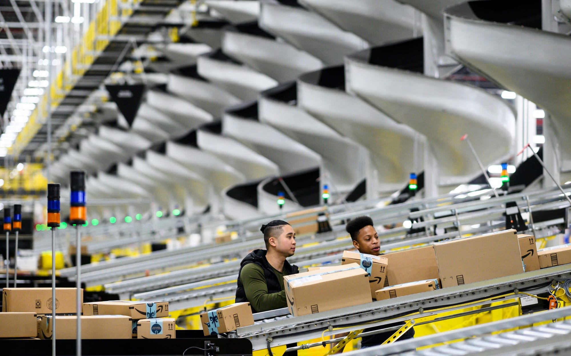 an Asian man with an undercut and a short ponytail and a Black man whose body is obscured check Amazon cardboard boxes on a c