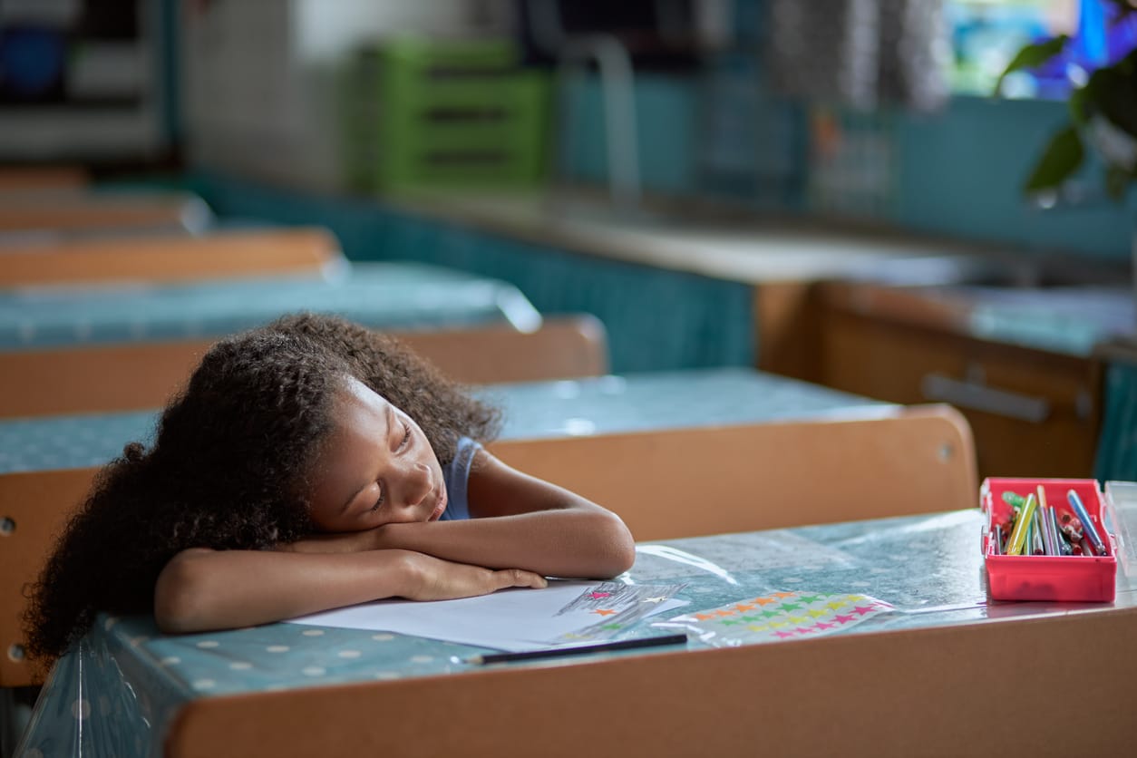 Shot of a young Black girl with curly hair in a ponytail sleeping at her desk in a classroom