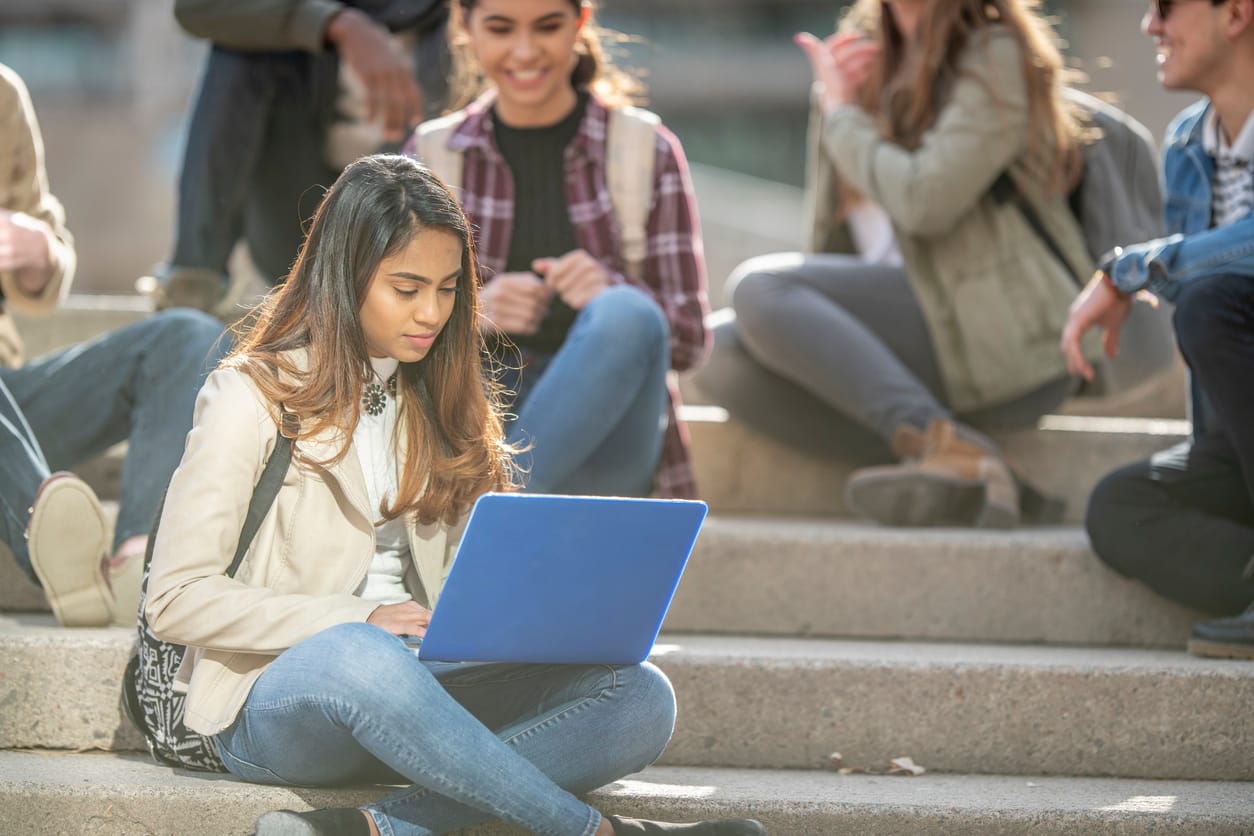 A small multi-ethnic group of University students sit outside on the steps together during break. They are each dressed causa