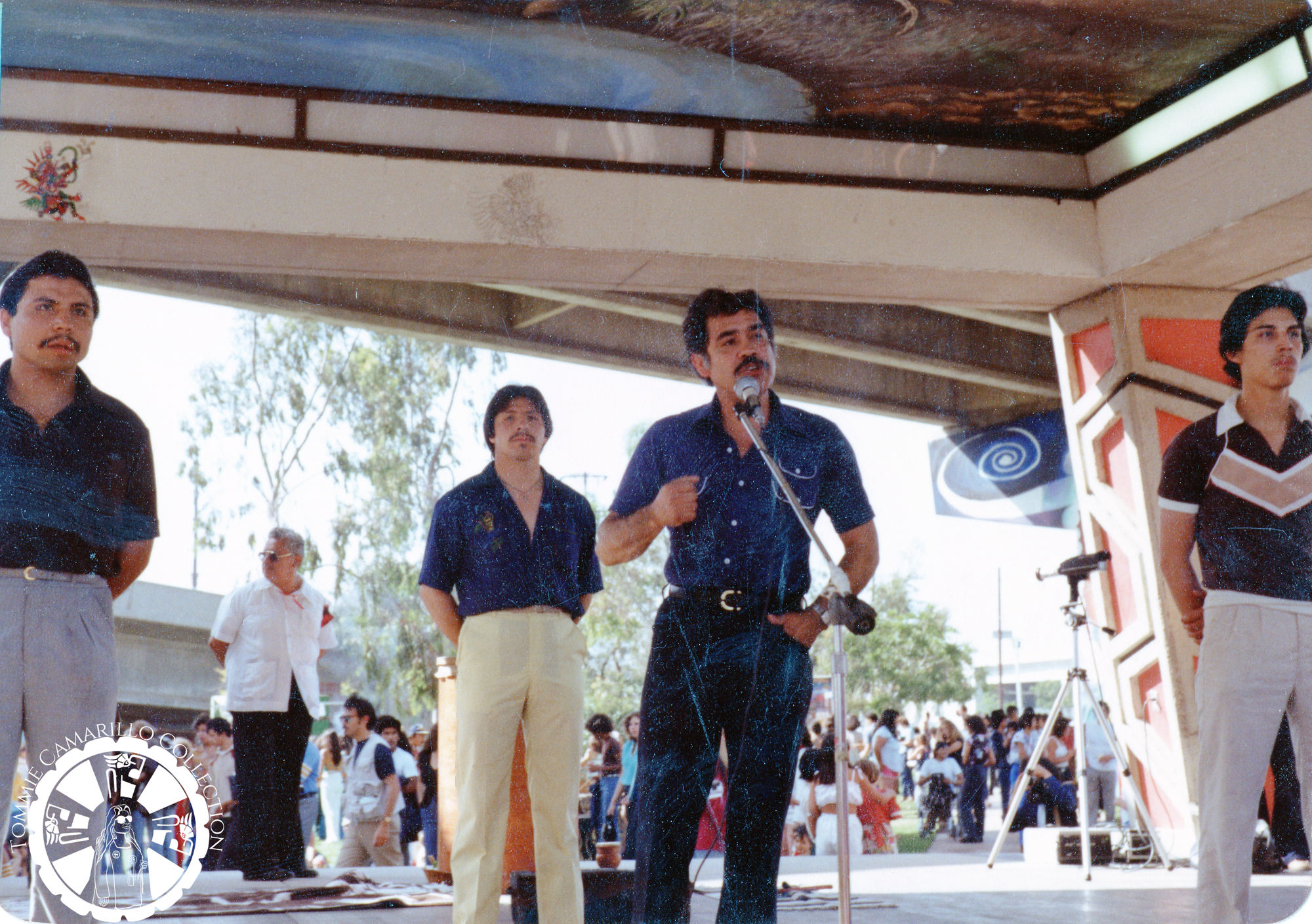 Chicano Civil Rights activist Corky Gonzalez stands in front of a microphone on stage with four other people who stand with their legs apart and hands behind their backs. Corky is wearing a blue button-down shirt and belted darker blue pants