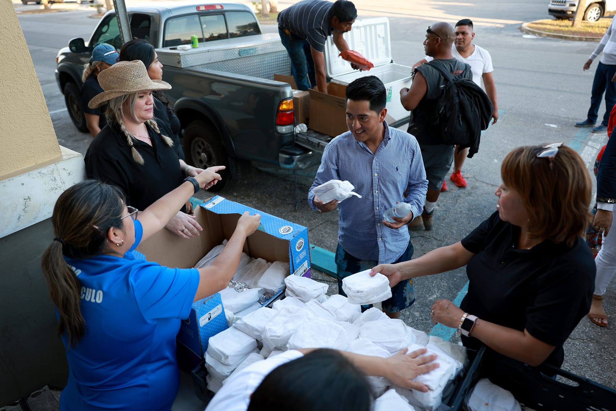 a man in a blue long sleeve button down holds a styrofoam takeout container as several people standing behind a folding table