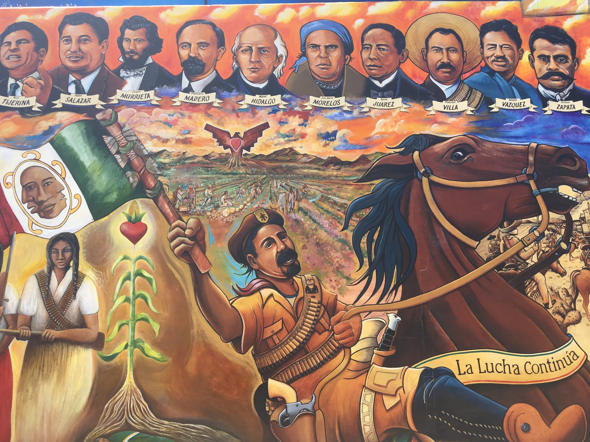 a close-up photograph of a colorful painted mural of a Brown Beret riding a brown horse and holding a Mexican flag. In the background of the mural is a field and mountains during sunset, and in the clouds are the painted busts of Tijerina, Salazar, Murrieta, Madero, Hidalgo, Morelos, Juarez, Villa, Vasquez, and Zapata