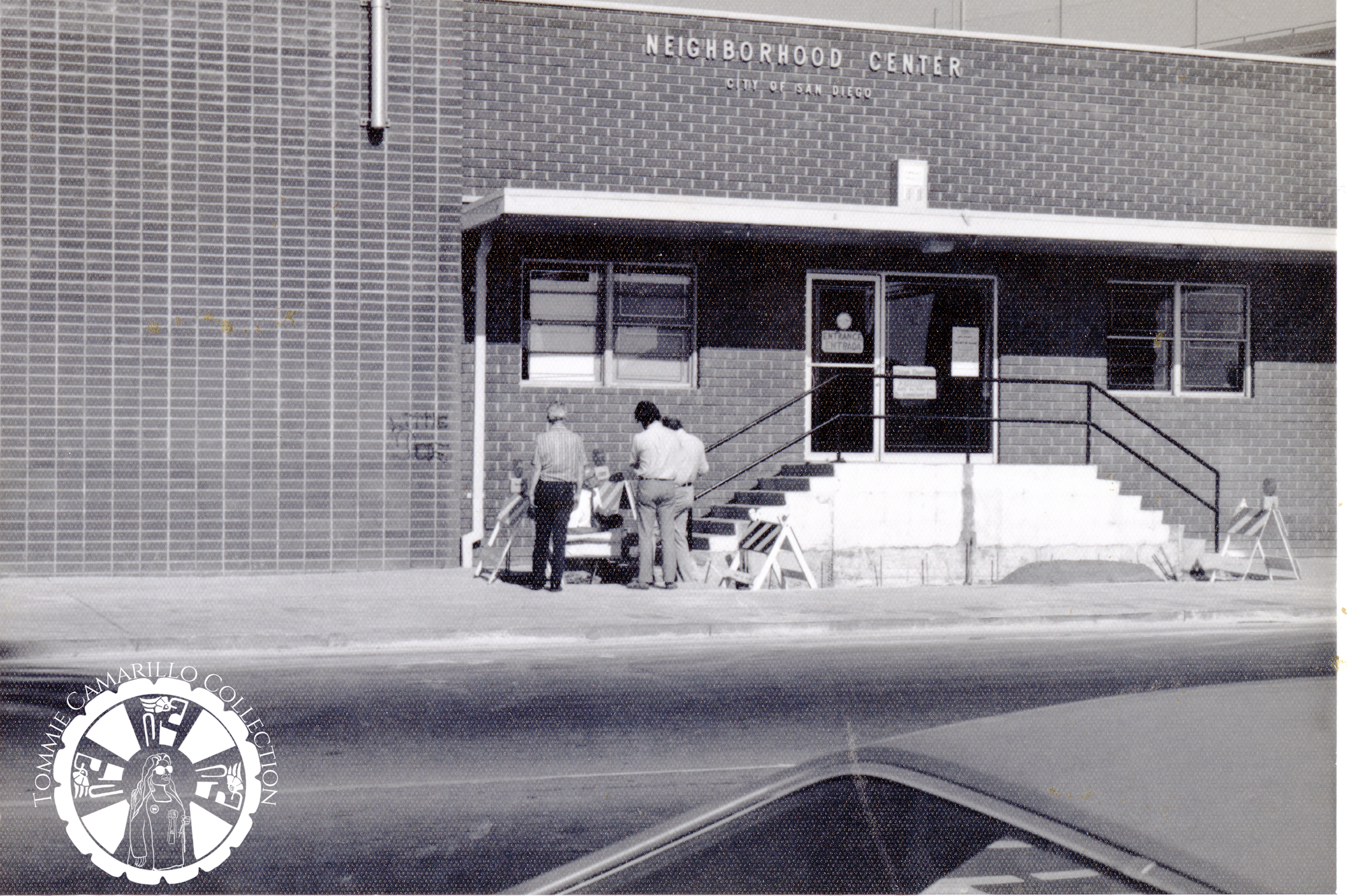 A black-and-white photograph of the exterior of a short brick building with white lettering above the door that reads "Neighborhood Center." Three people in long pants and short sleeve button-downs stand at the base of the short staircase leading up to the door 