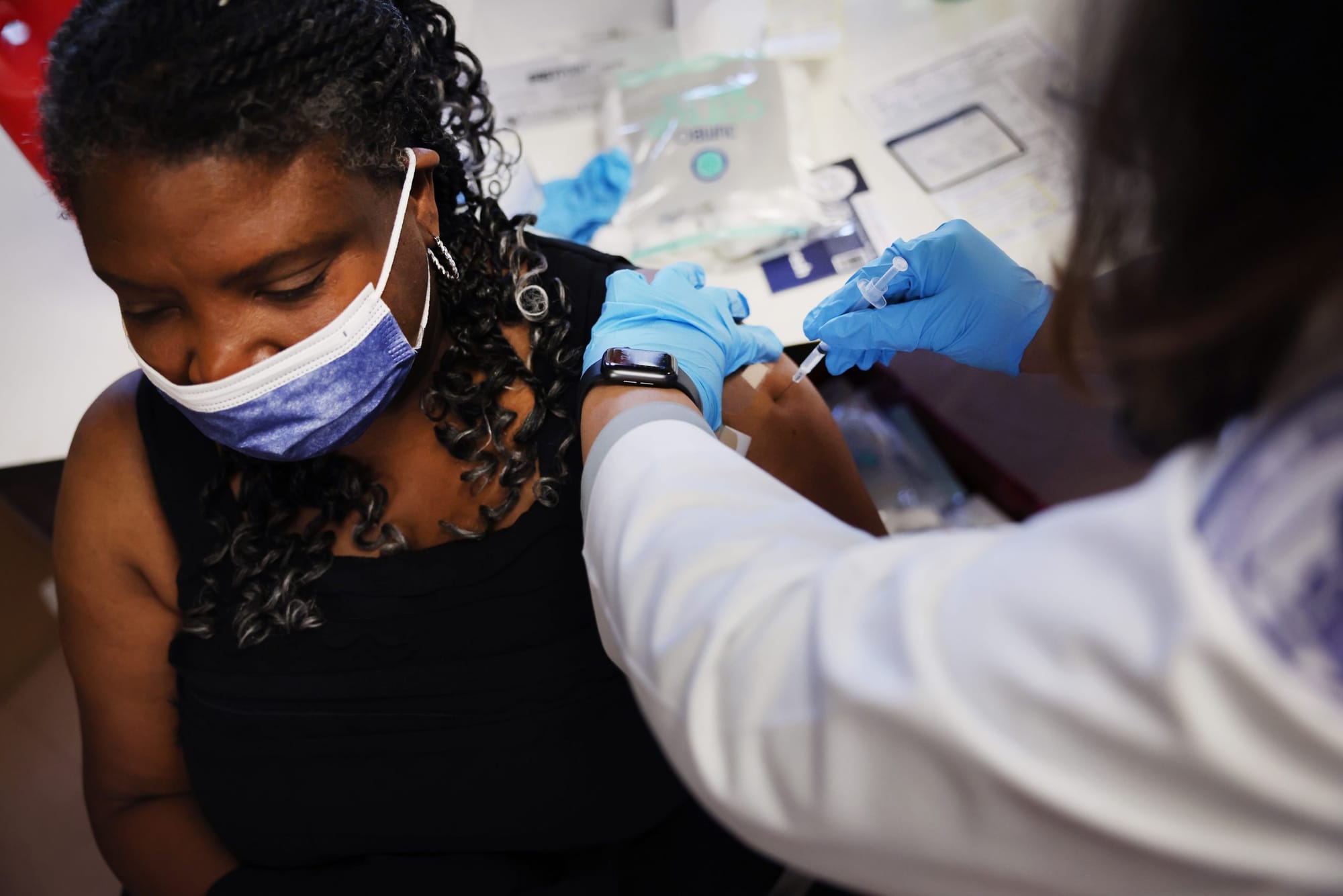 color photograph of a black woman sitting in a chair as she receives an Omicron booster. she wears a blue medical mask