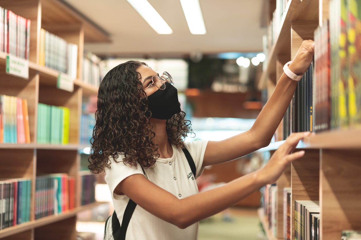 color photograph of a young teenage girl wearing a black mask, white t-shirt, and backpack, looking through a school library
