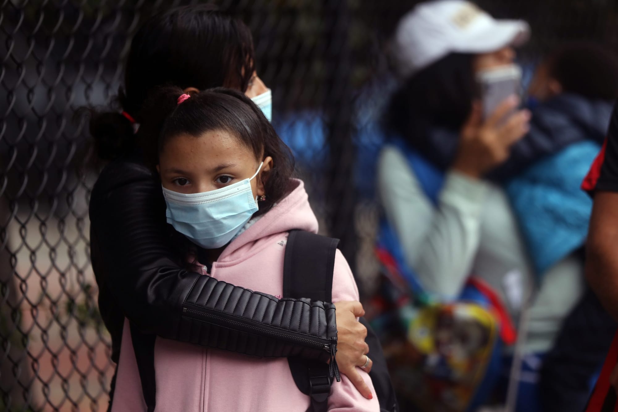 color photograph of a young black girl wearing a backpack and light pink hoodie with a blue medical face mask standing in fro