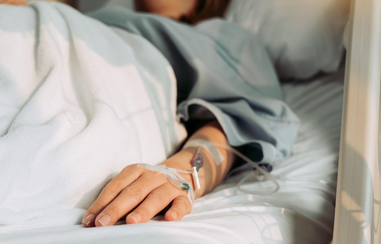 color stock photo of a woman's hand with an IV drip hooked up. her face is slightly out of frame as she lies in a hospital be