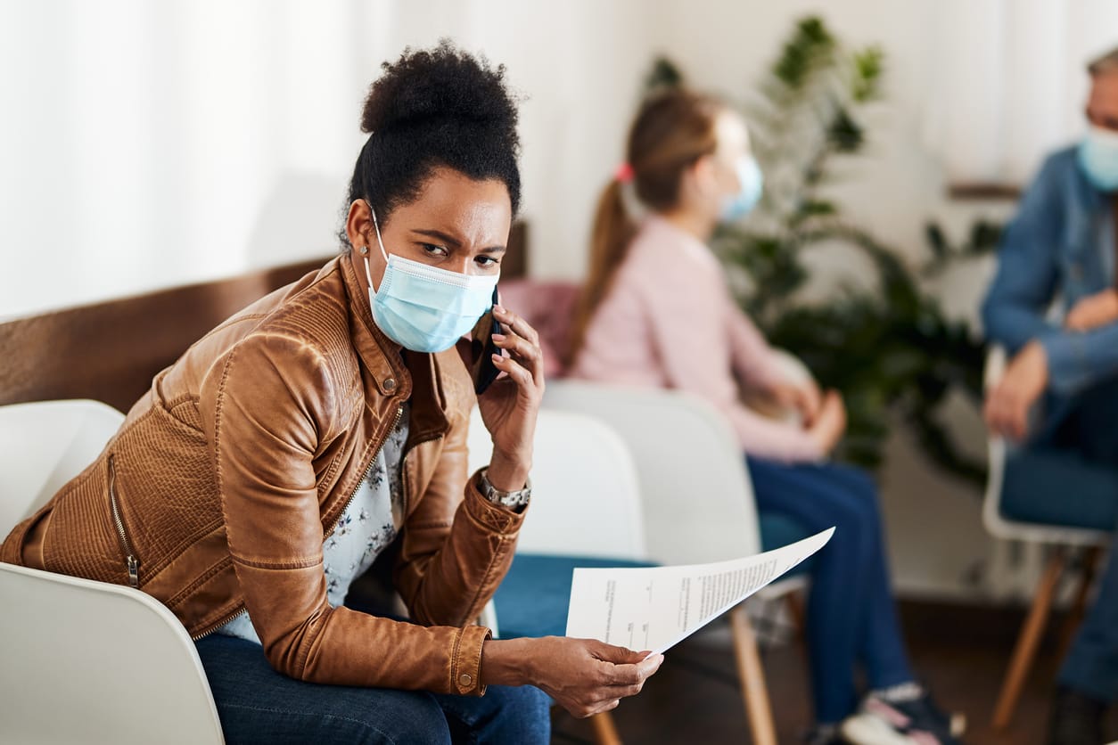 stock color photo of an African American woman communicating over cell phone while wearing face mask and waiting for medical