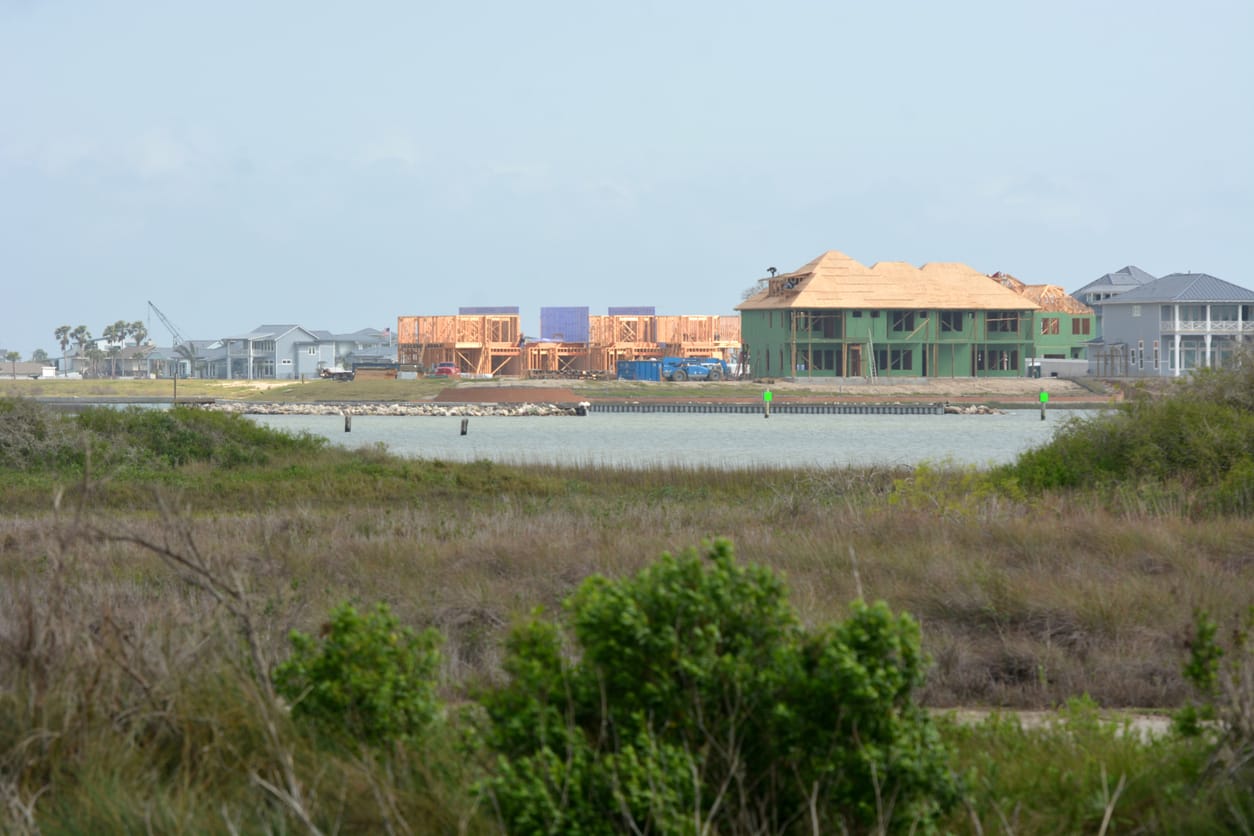 color photo of grassy wetlands in the foreground, a strip of water in the midground, and unfinished construction of large hou