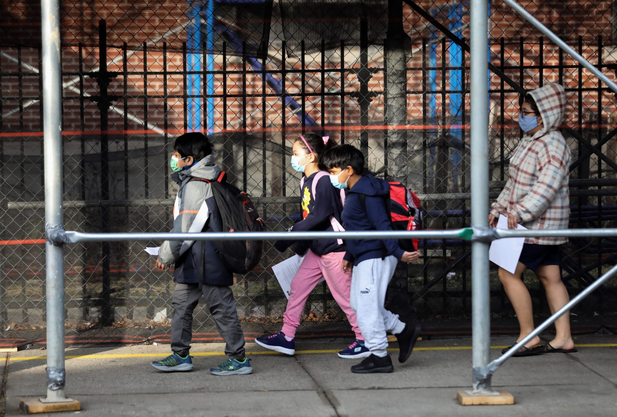 color photograph of three young children wearing long pants, jackets, and face masks walking under scaffolding in New York Ci