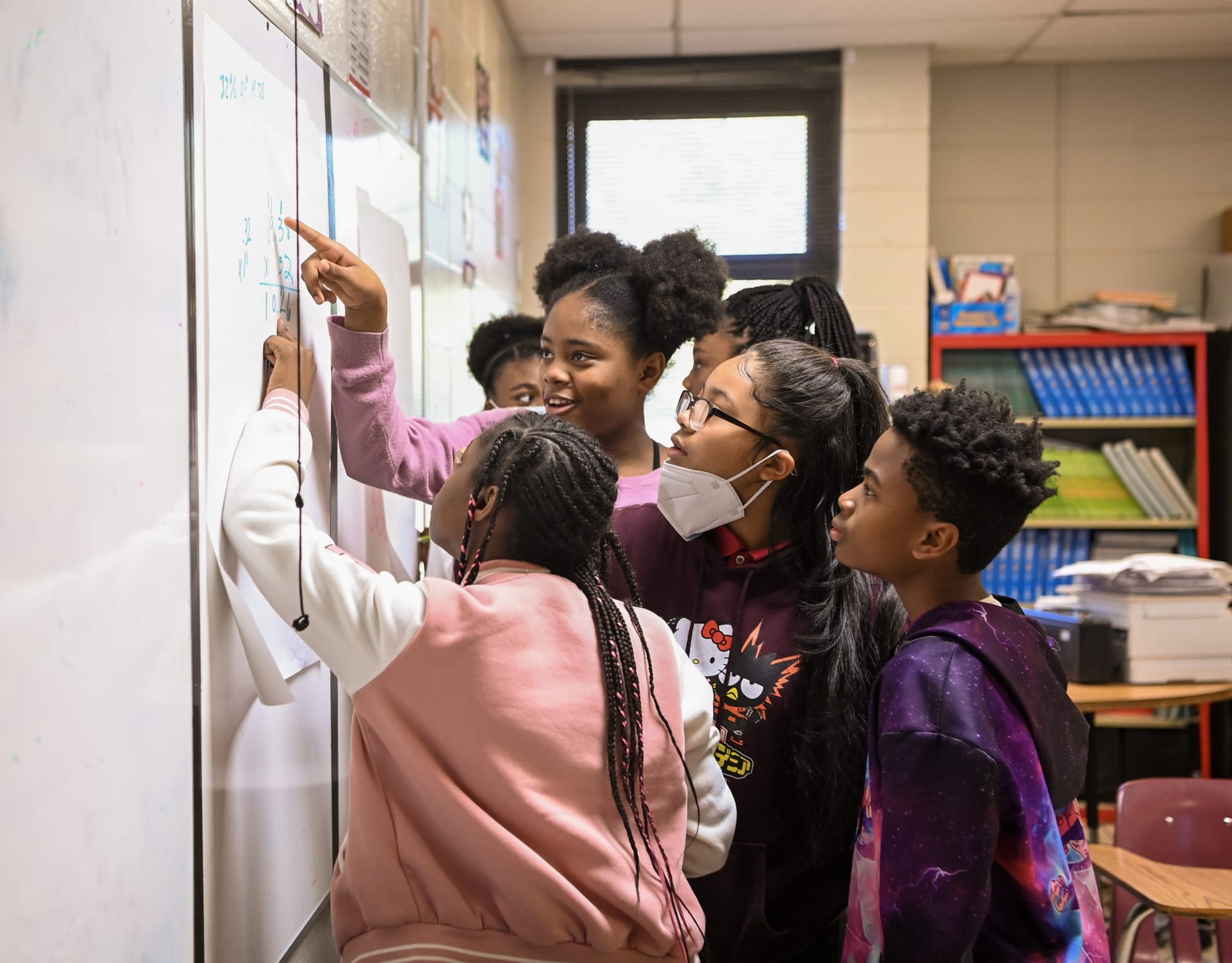 color photograph of four elementary school students gathered at the white board. They point to the board as they discuss a ma