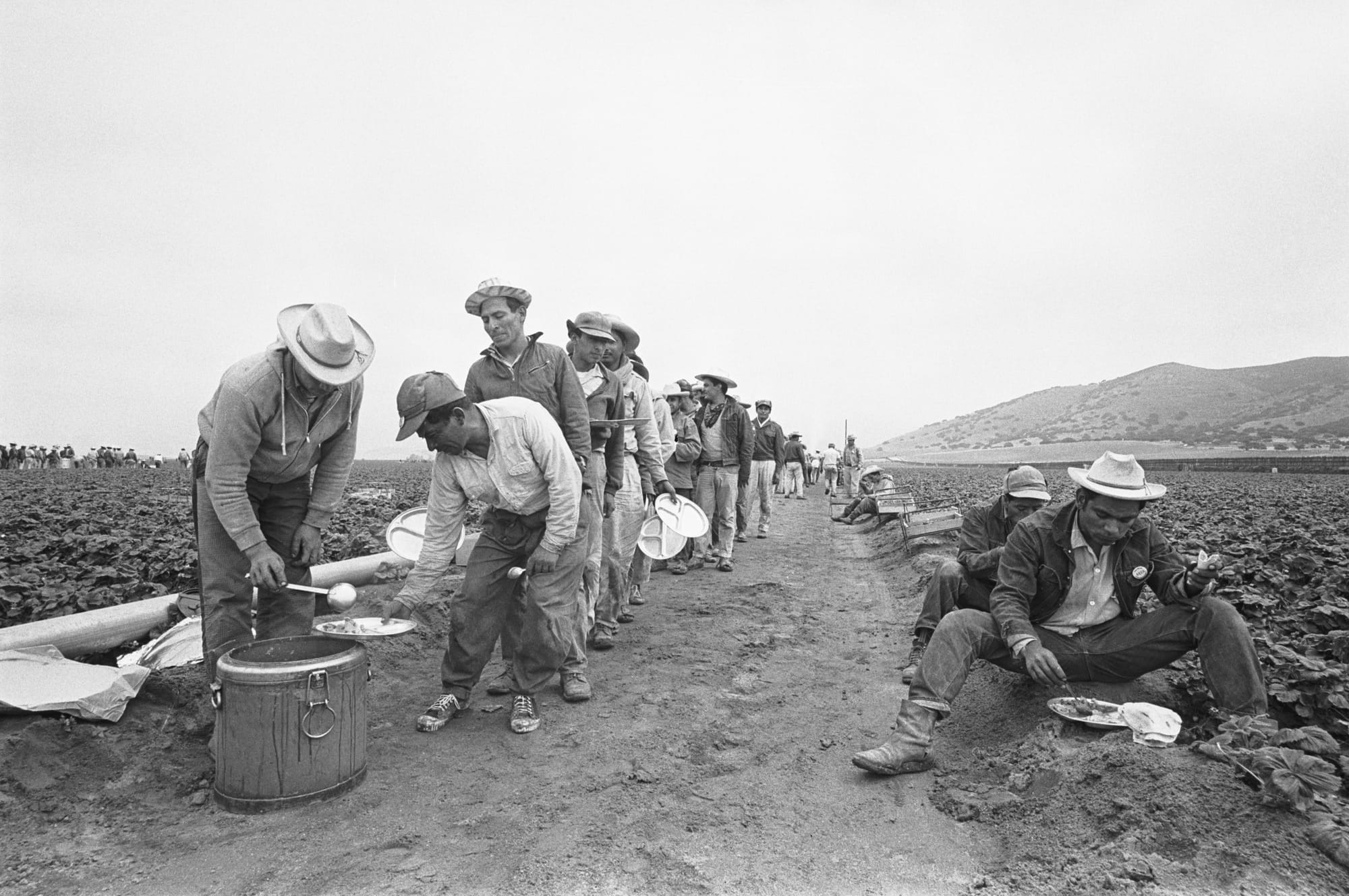 black-and-white historical photo of braceros, latino farmworkers, standing and sitting on the side of a dirt road