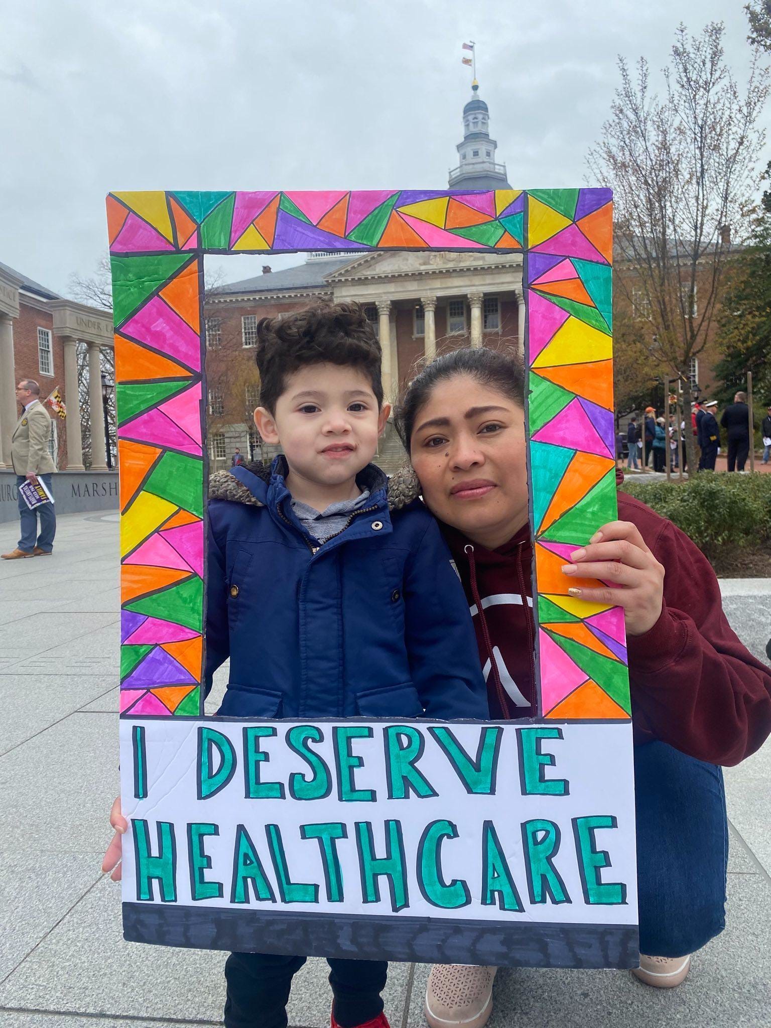color photograph of a Latinx woman with her hair tied back holding up a colorful paper frame around her and her son with the text "I deserve healthcare" in all caps teal lettering at the bottom