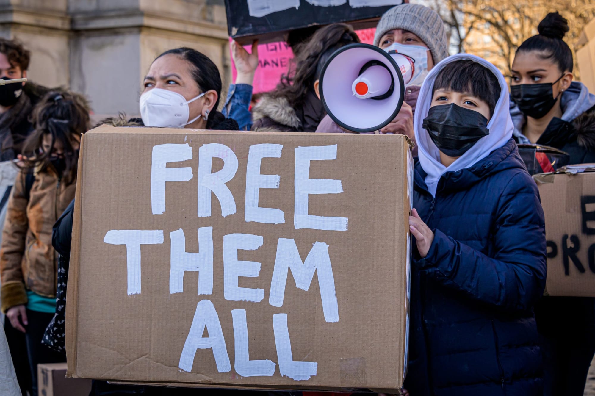 color photograph of an outdoor protest. two people hold a large cardboard sign with white handwritten text that reads "Free t