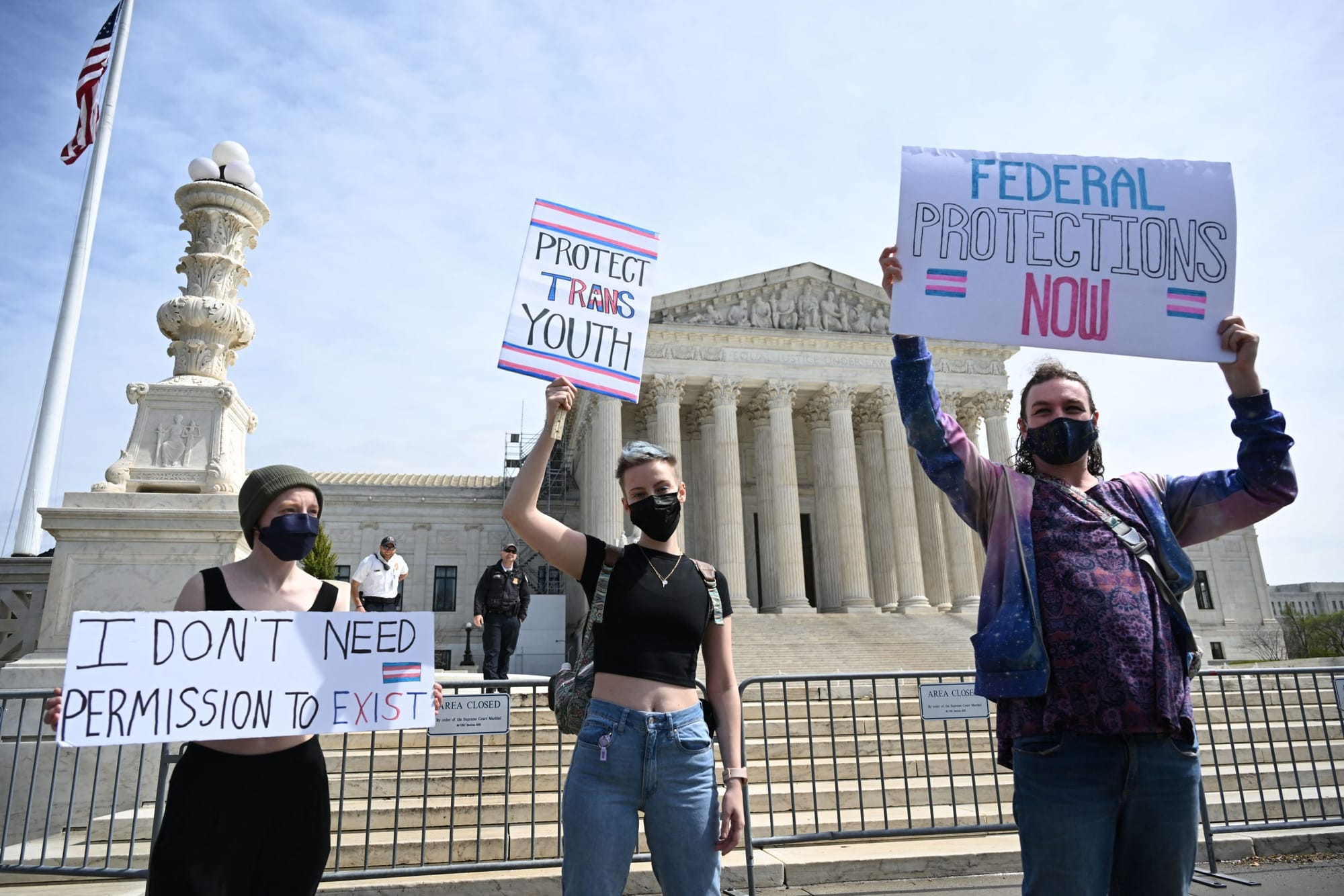color photograph of three protesters outside the U.S. Supreme Court building. they hold white posters with trans flags and pi