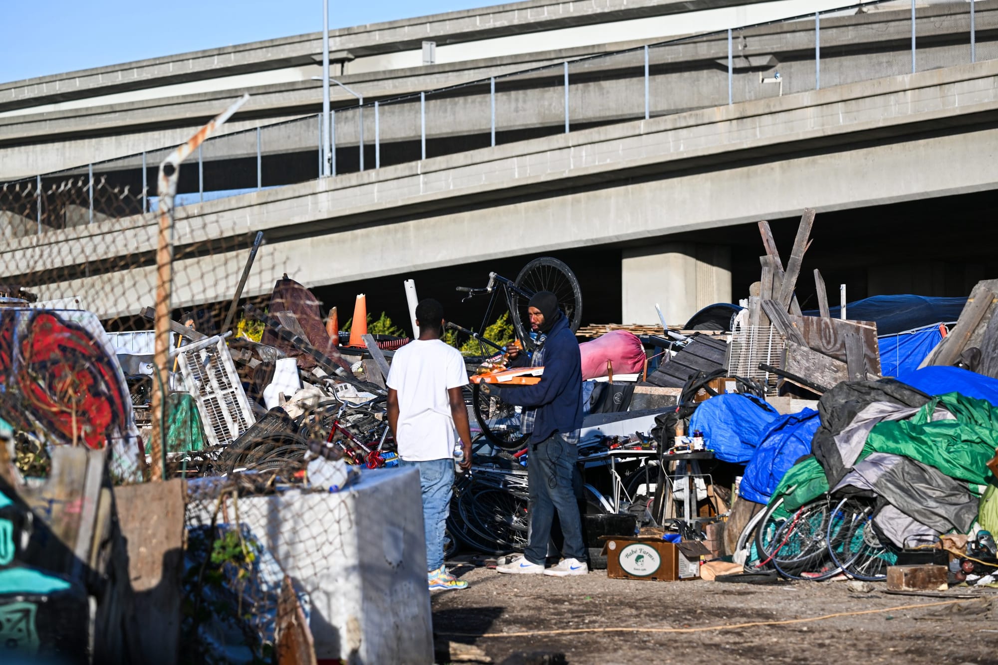 color photograph of two Black men standing in the midground in a homeless encampment near a highway ramp. Tarps, bicycles, pl