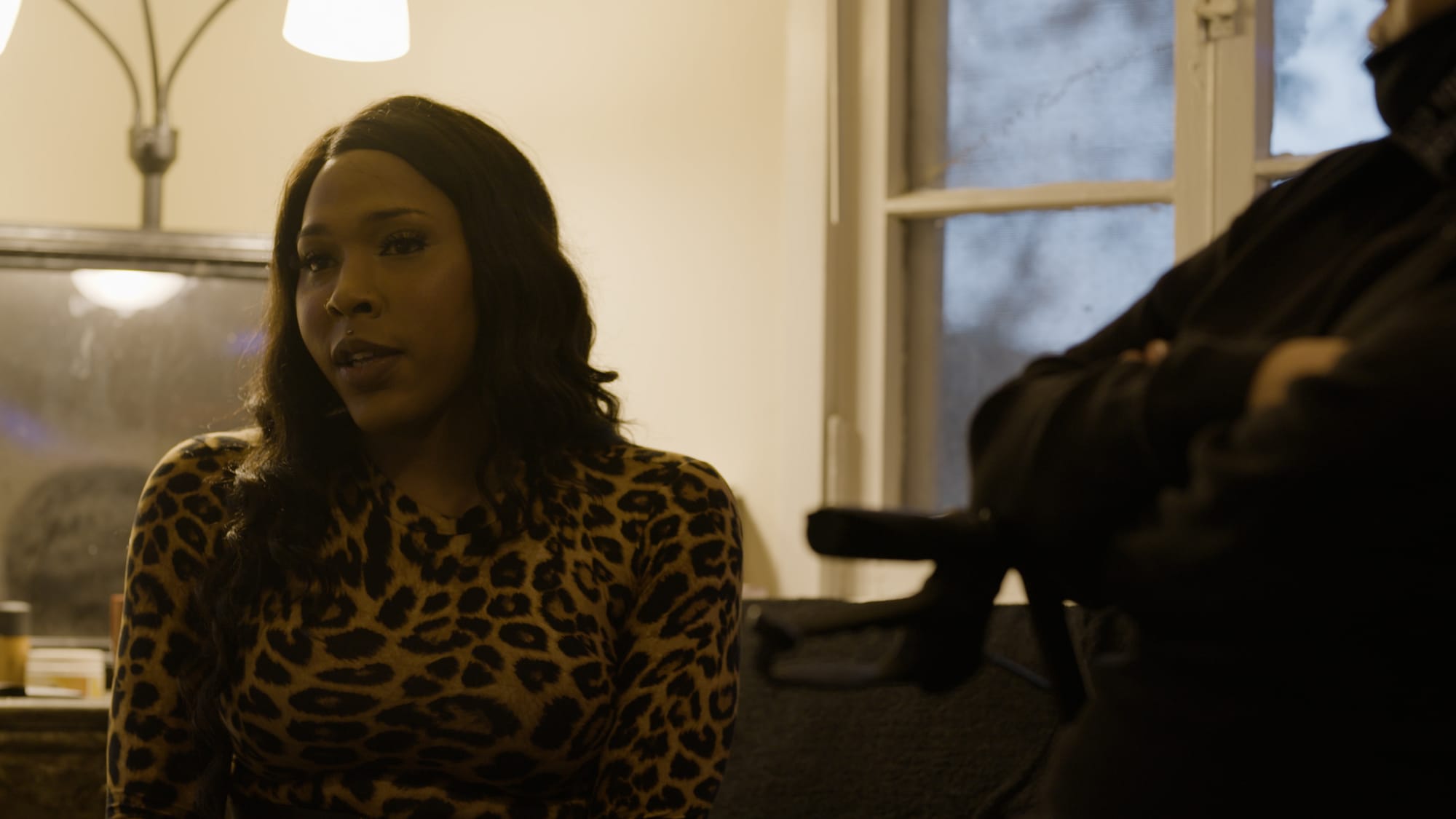 color photograph of a Black trans woman with shoulder-length black hair sitting in a dark living room back-lit by a standing lamp. She looks to the left of the frame as she talks