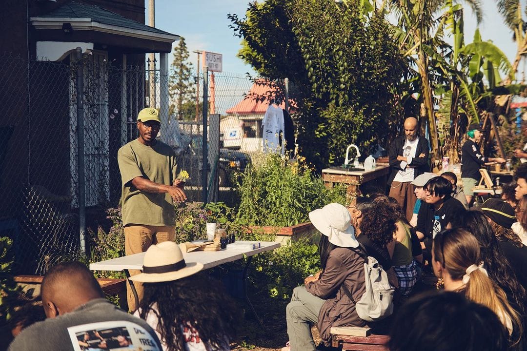 color photograph of a Black man facing a group of about 20 people sitting with their sides or backs to the camera in a community garden