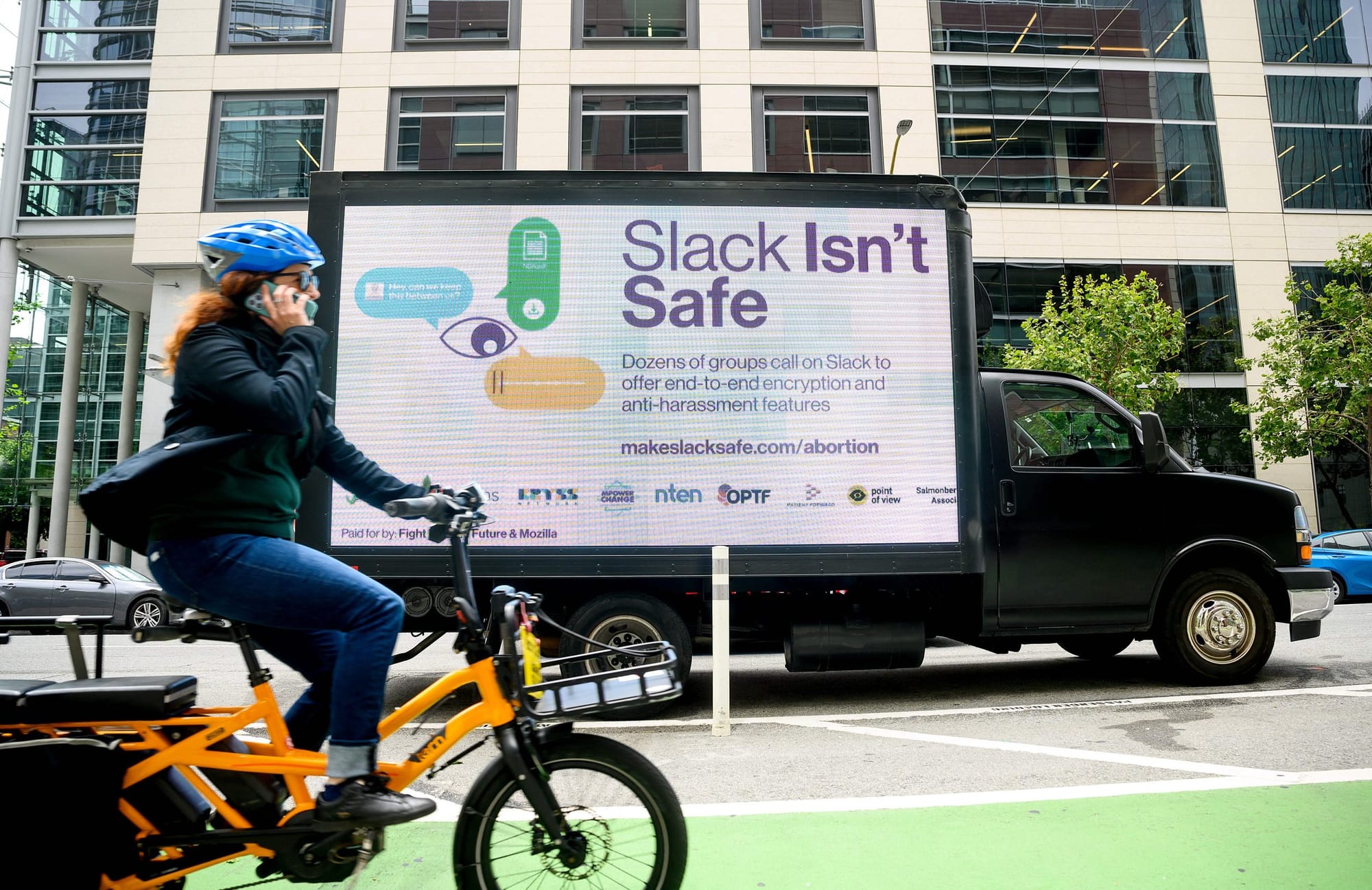 color photograph of a person riding an orange bicycle in a bike lane past a mobile digital billboard that reads "slack isn't
