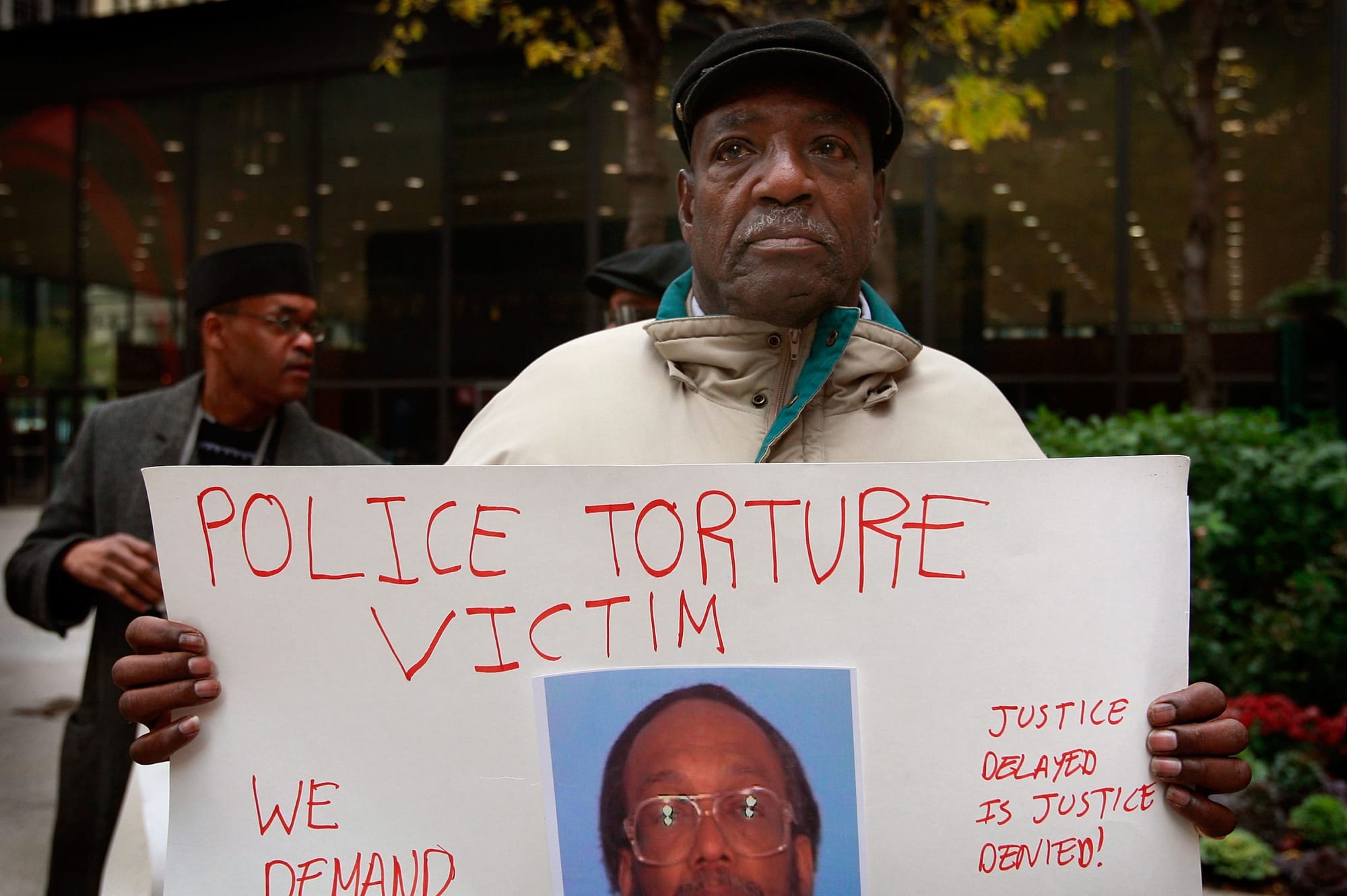 color photograph of a black man holding a large white poster with red text reading "police torture victim" and "justice delay