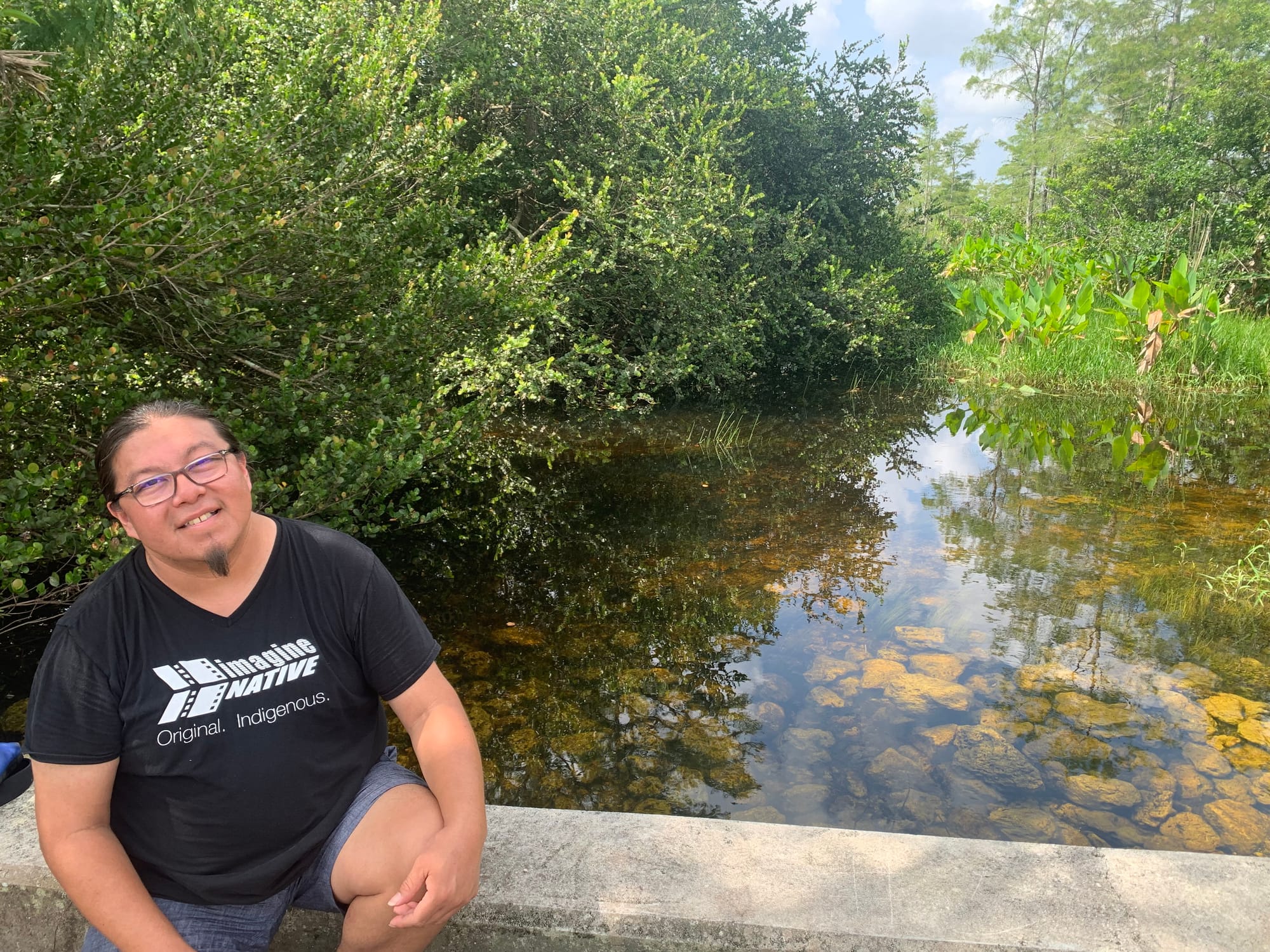 color photograph of an Indigenous person in a black t-shirt that has white text reading "imagine native. original. indigenous." they sit on a stone railing in front of clear shallow water through which stones are visible. dense trees and foliage line the body of water