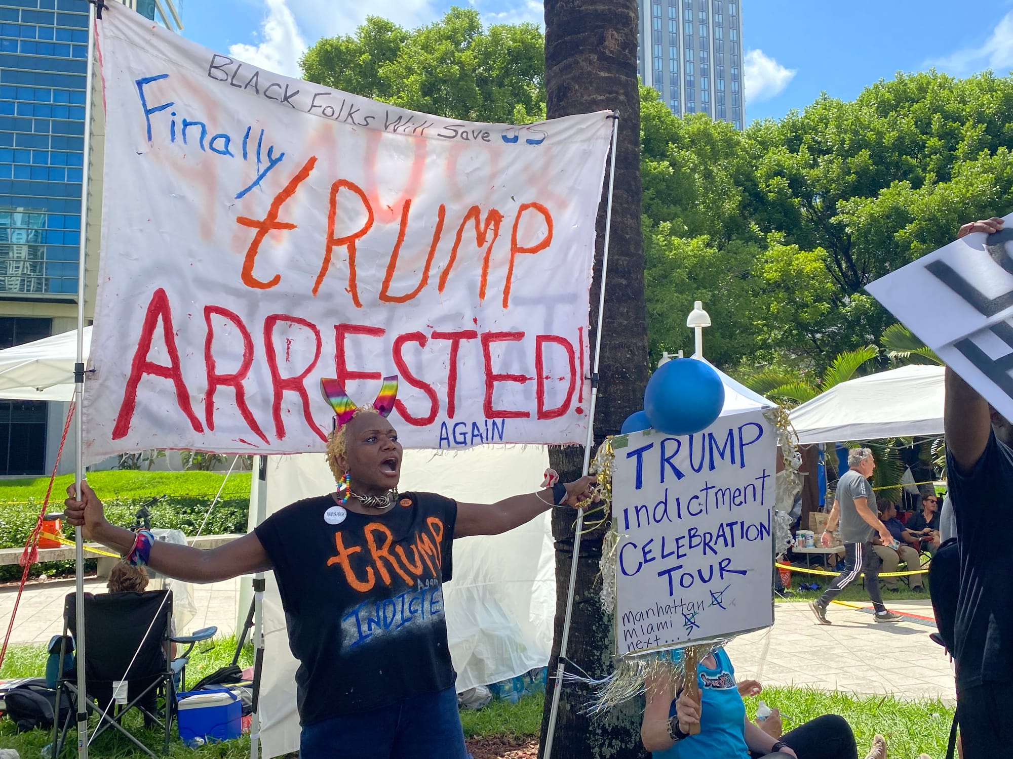 color photograph of a Black woman with short blonde hair holding up a large banner on two sticks that reads "Black Folks Will Save Us, Finally Trump Arrested! Again"