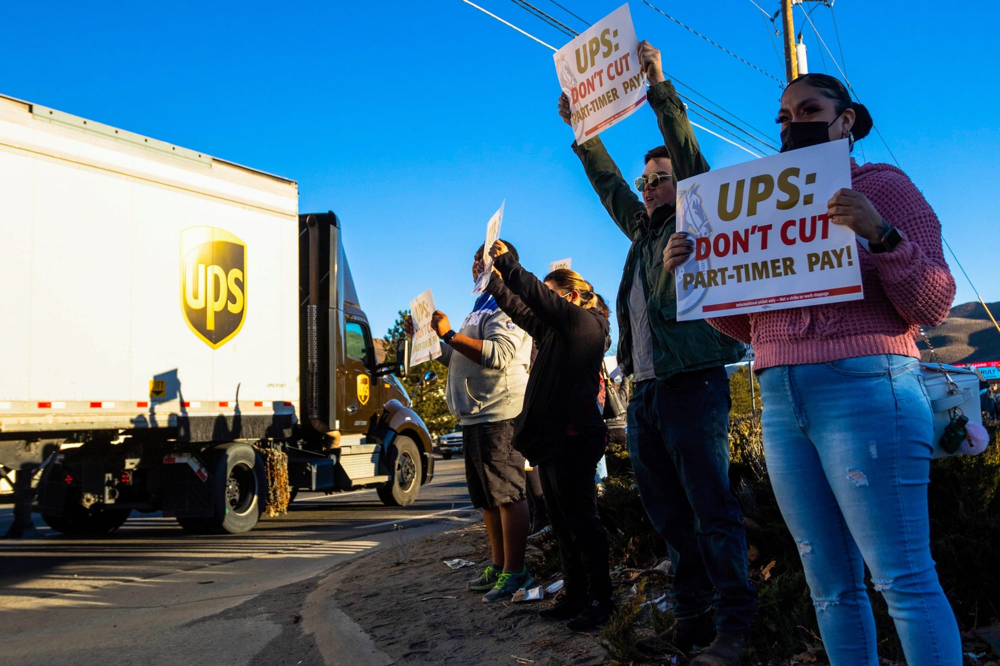 color photograph of a small protest of UPS workers standing on the right side of the frame holding posters that read "ups: do
