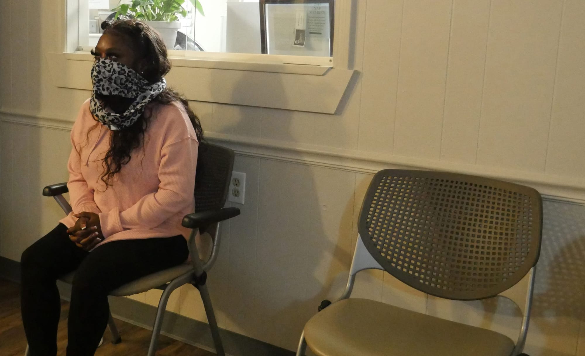 color photograph of a Black woman wearing a patterned scarf over her mouth and nose. she sits with her hands in her laps agai