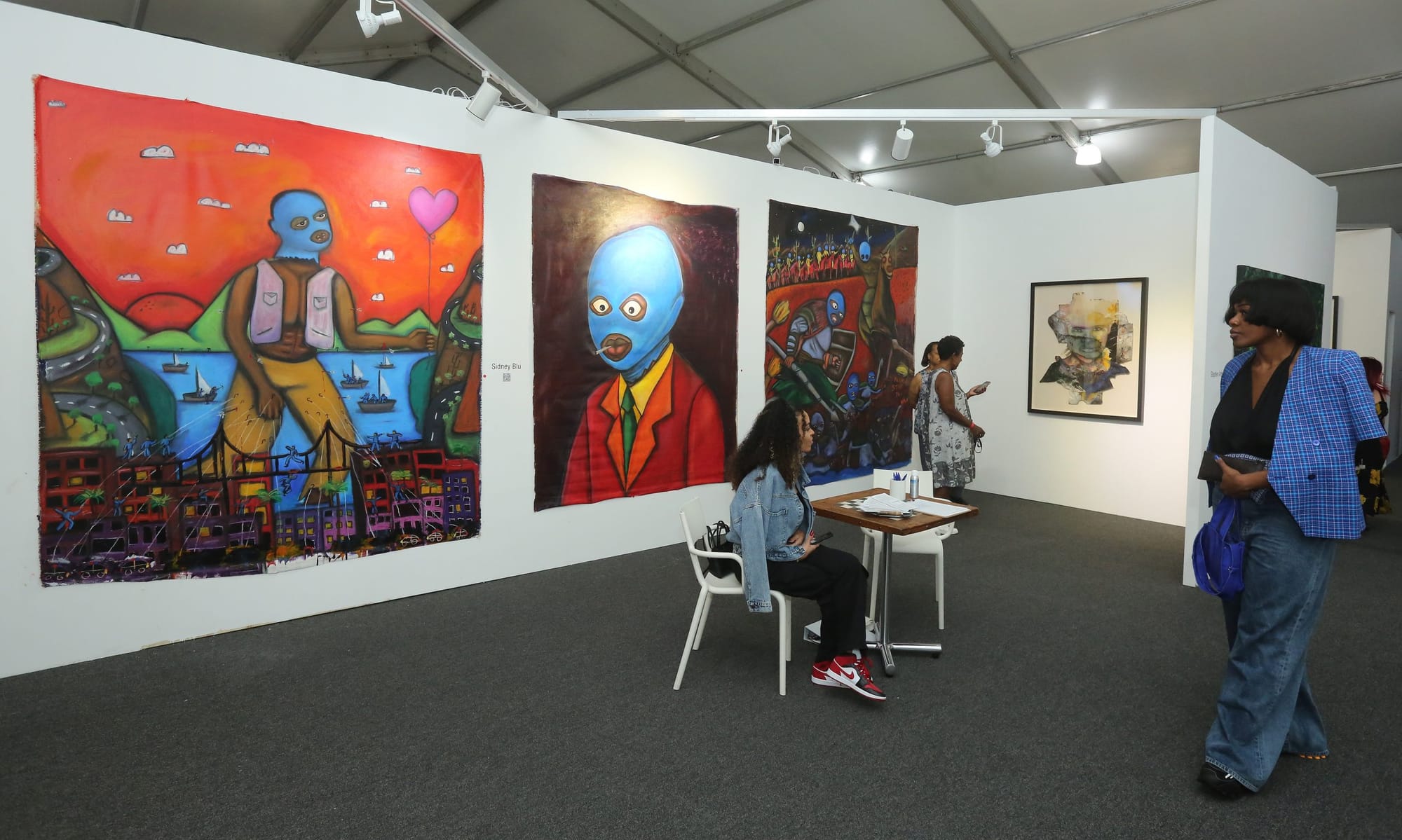 color photograph of an indoor art exhibit. large paintings are hung on a white wall. the paintings use vivid reds and darker