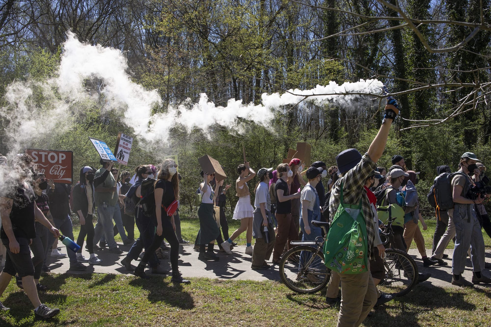 color photograph of an outdoor protest against cop city in the woods. a line of protesters walk from left to right in the fra