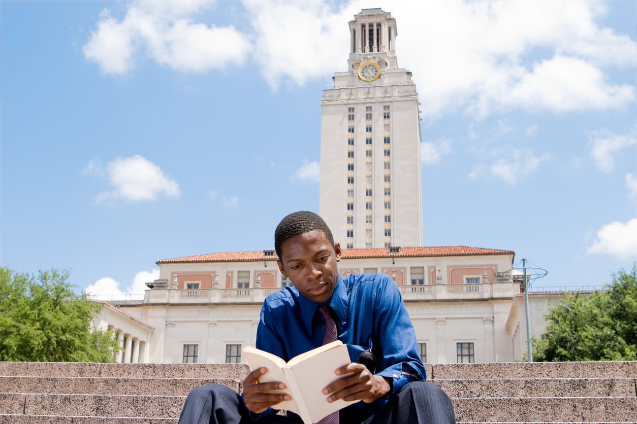 color stock photo outside of a Black college student wearing blue button-down shirt. he has close-cut shaved hair and reads a